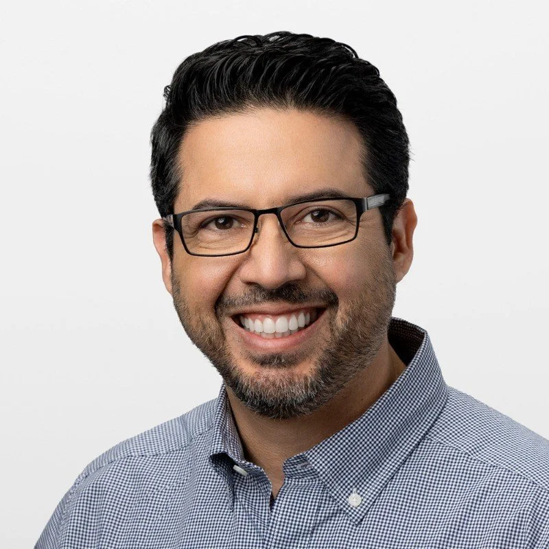 Headshot of a smiling man with dark hair, glasses, and a beard, wearing a collared shirt against a plain white background.
