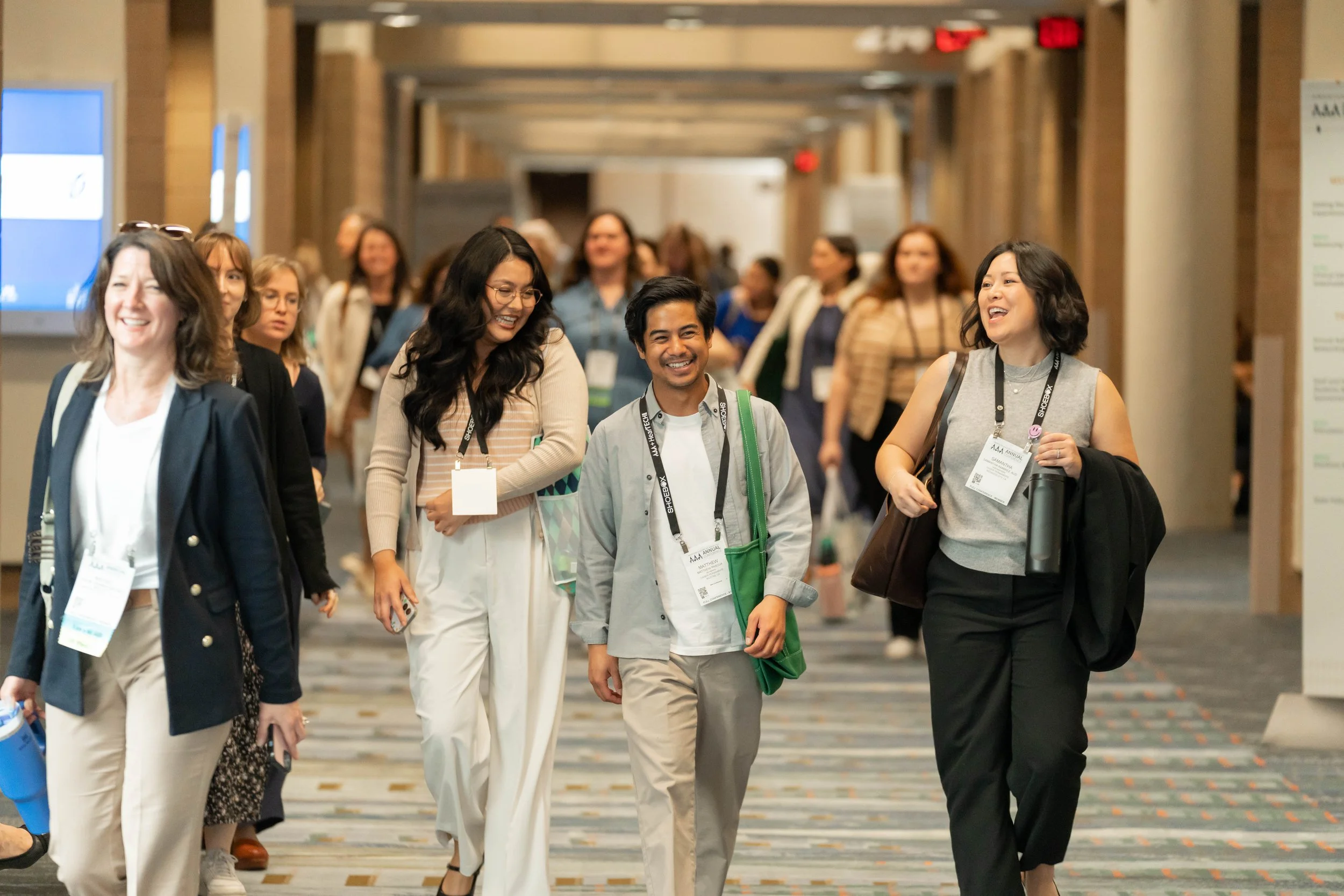 Conference attendees walking and smiling in a hotel corridor, wearing name tags and carrying bags.
