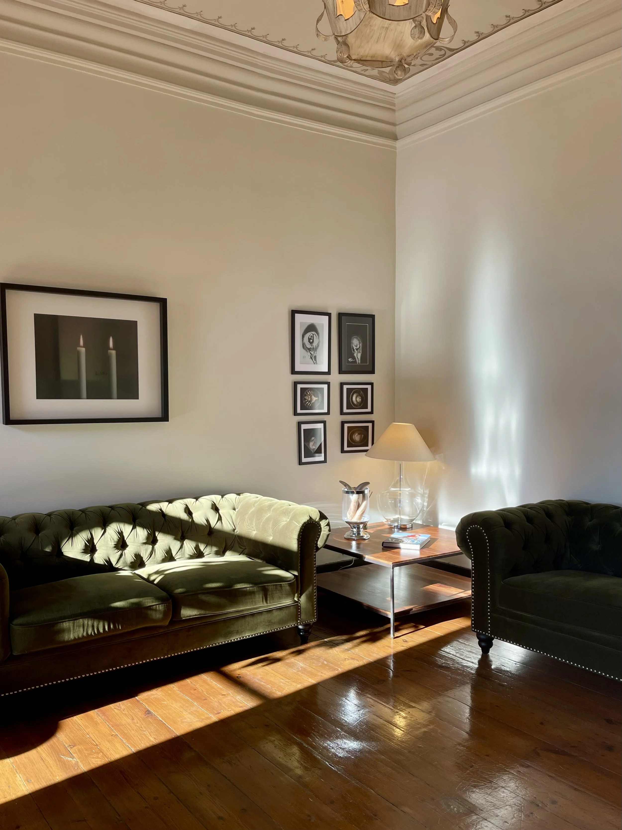 A living room with two green tufted sofas, a wooden side table, a lamp, and framed black-and-white artwork on the wall, with sunlight streaming onto the polished wooden floor.