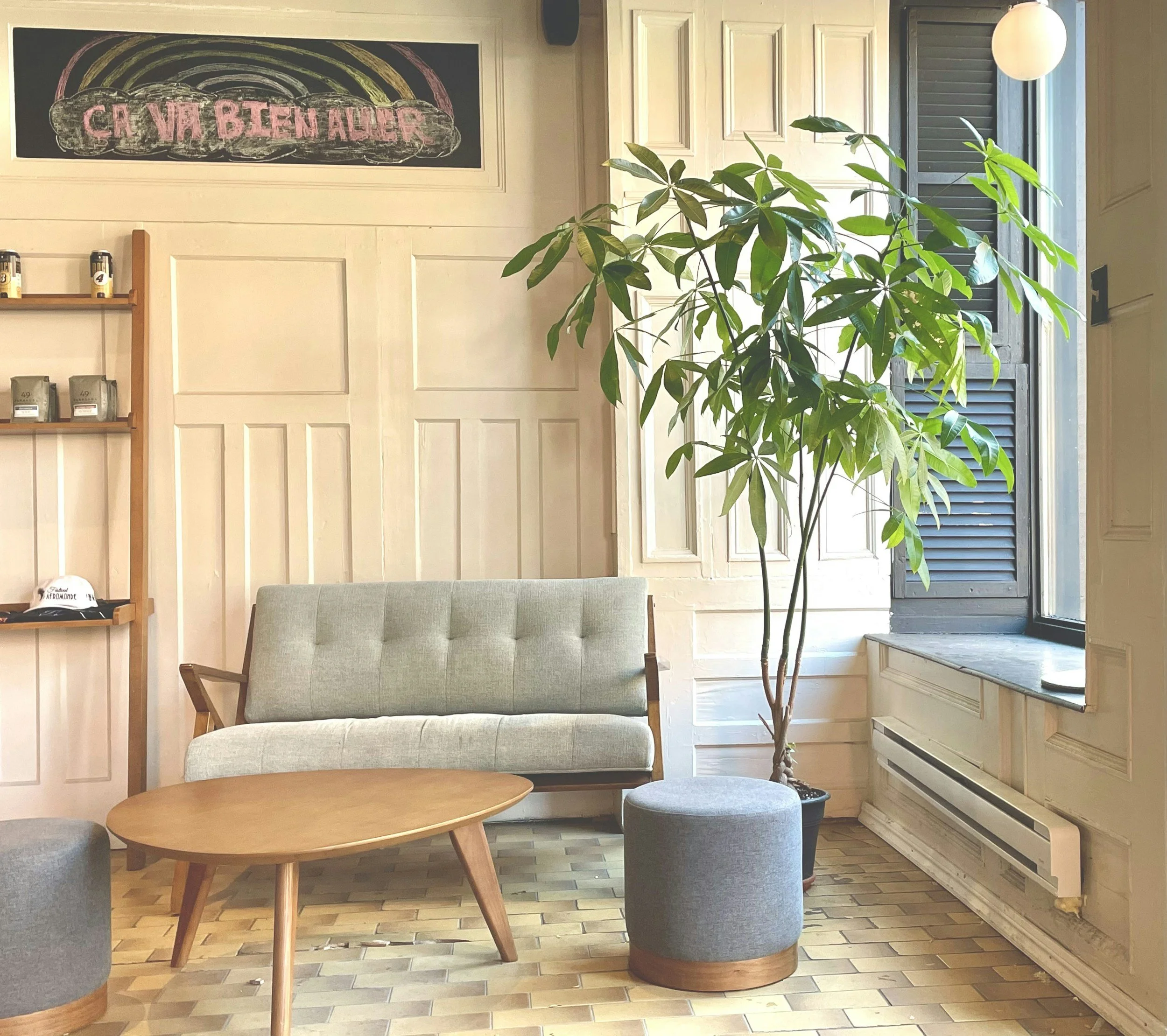 A cozy living room corner with a light green upholstered sofa, a wooden coffee table, a round gray ottoman, and a large potted plant near a window. There is a wooden shelf with jars and a hat, cream paneled walls, and a blackboard sign with French writing at the top.
