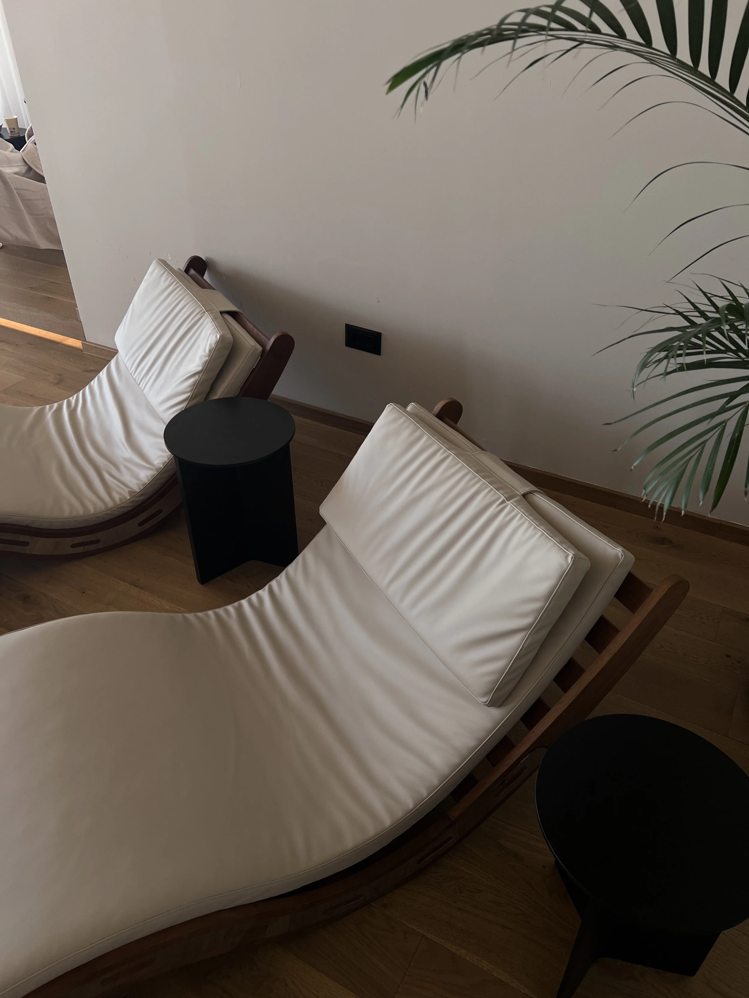 Two modern lounge chairs with cream-colored cushions and wooden frames, separated by a small black round side table, in a minimalist room with wooden flooring and a white wall.