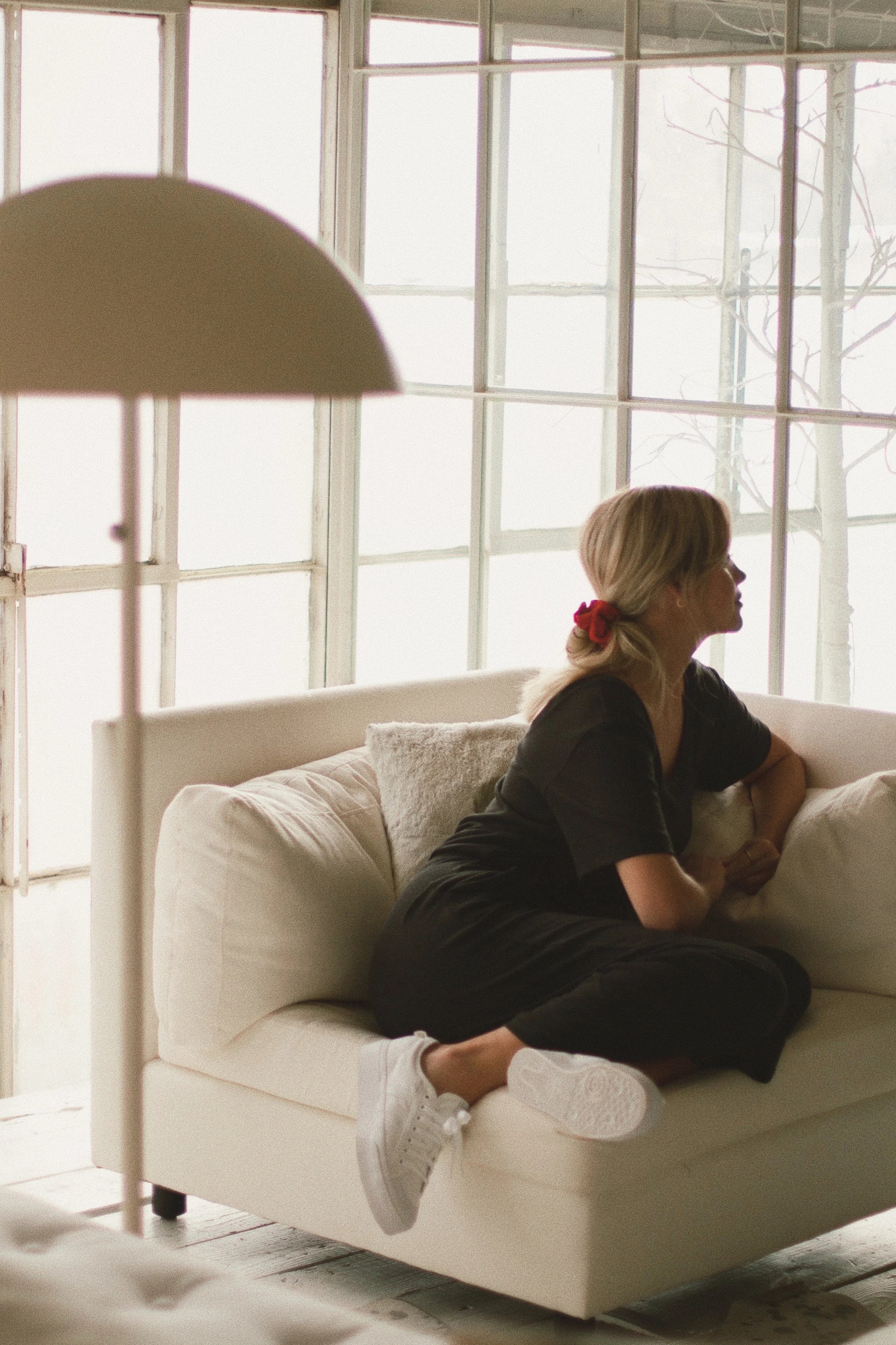 A woman with blonde hair tied back with a red ribbon, wearing black clothes and white sneakers, sitting on a white sofa in a room with large windows, looking to the right.
