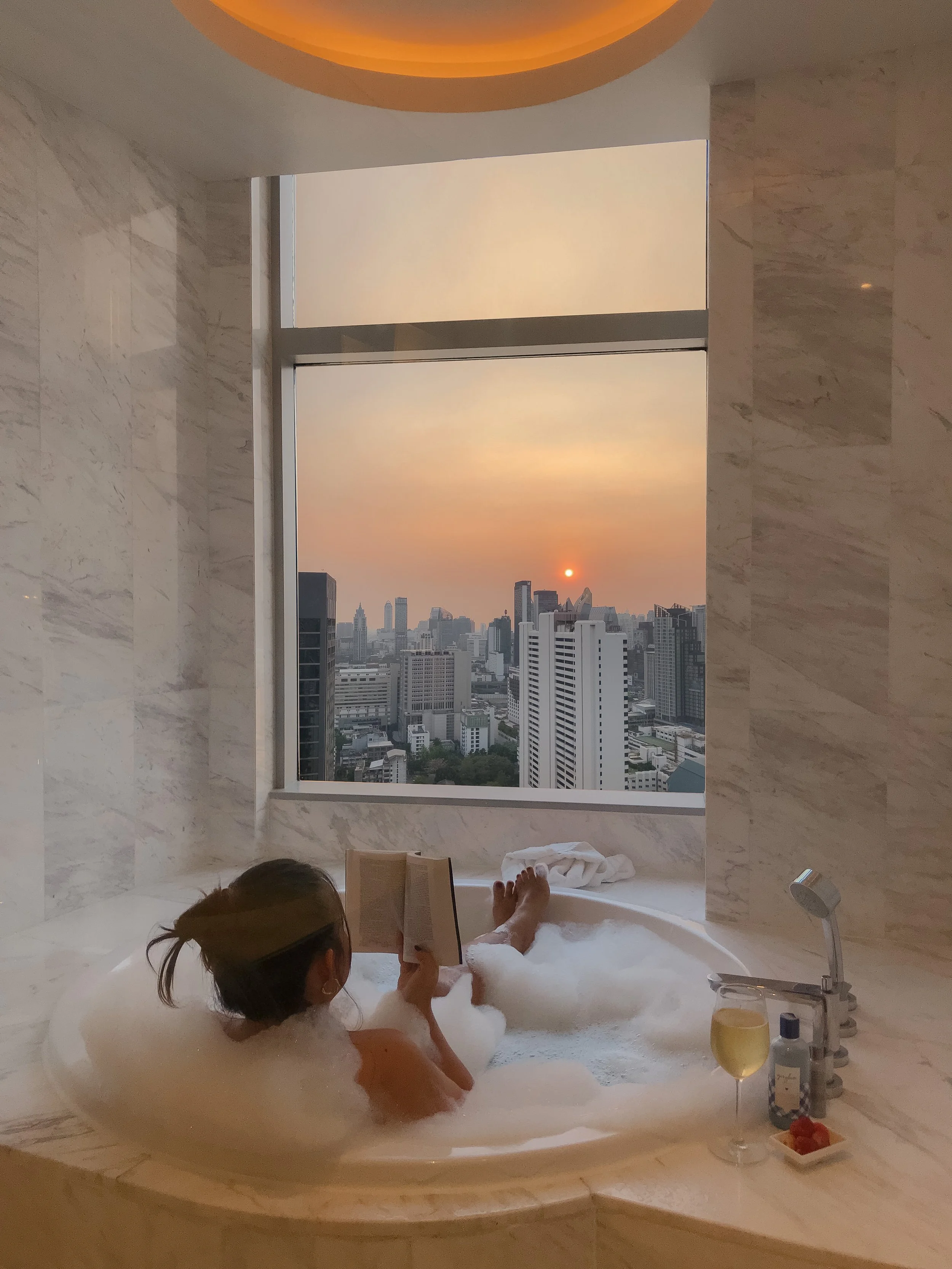 A woman relaxing in a bubble bath in a high-rise hotel bathroom, reading a book with a city skyline view at sunset outside the window, with a glass of white wine and bath amenities on the edge of the tub.