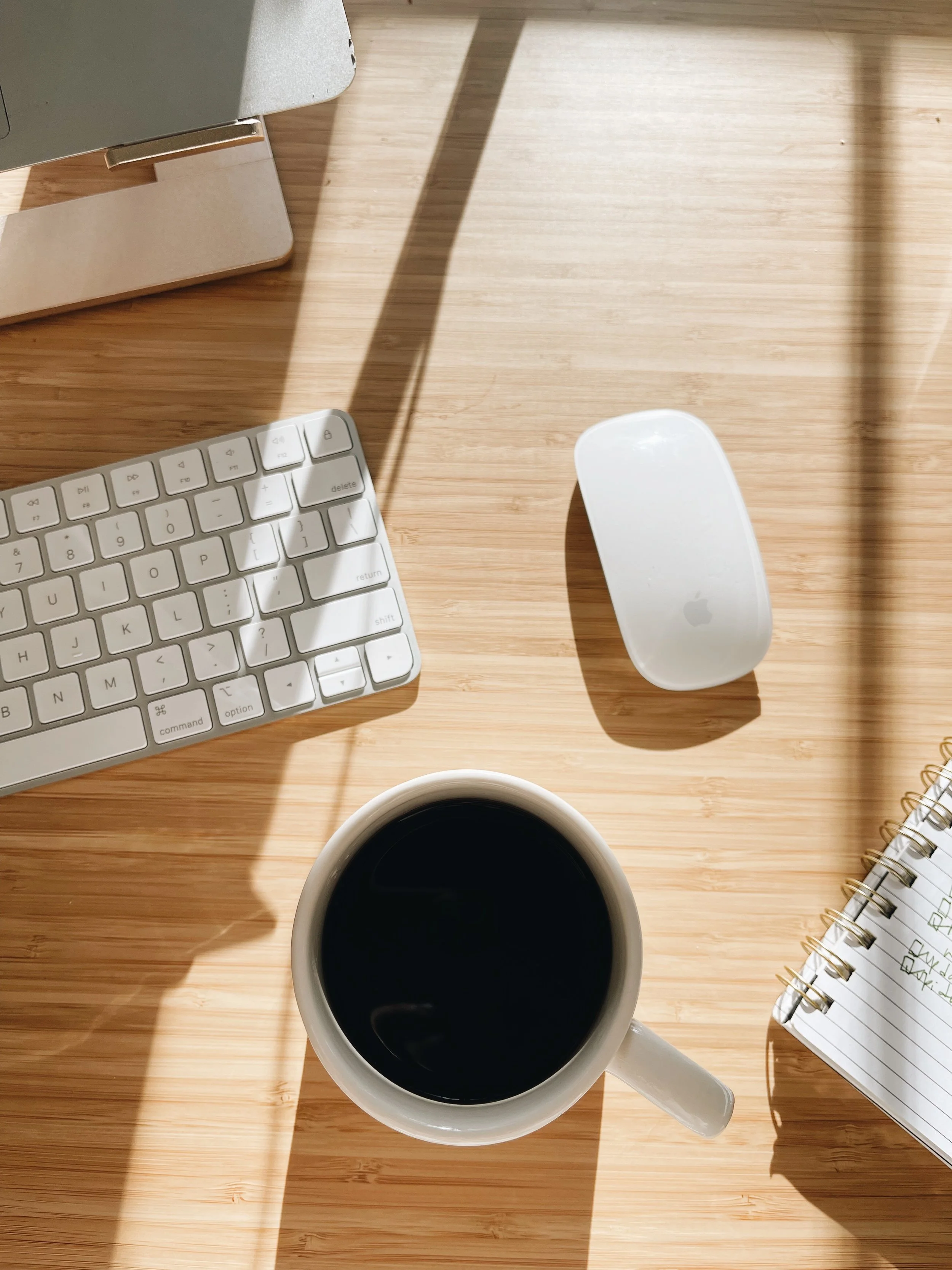 A wooden desk with a computer keyboard, an Apple Magic Mouse, a cup of black coffee, a notepad with handwritten notes, and part of a laptop or monitor in the upper left corner, with sunlight casting shadows.