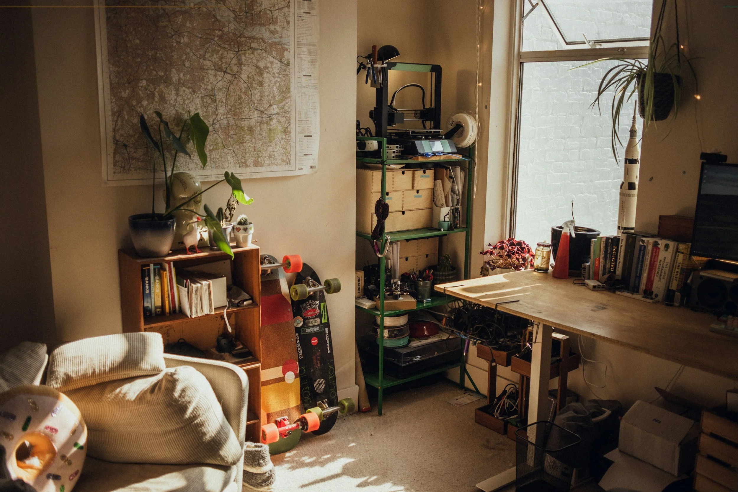 A cozy room with a large map on the wall, a skateboard leaning against a small bookshelf, a desk next to a window with books and a model rocket, and various plants and household items.