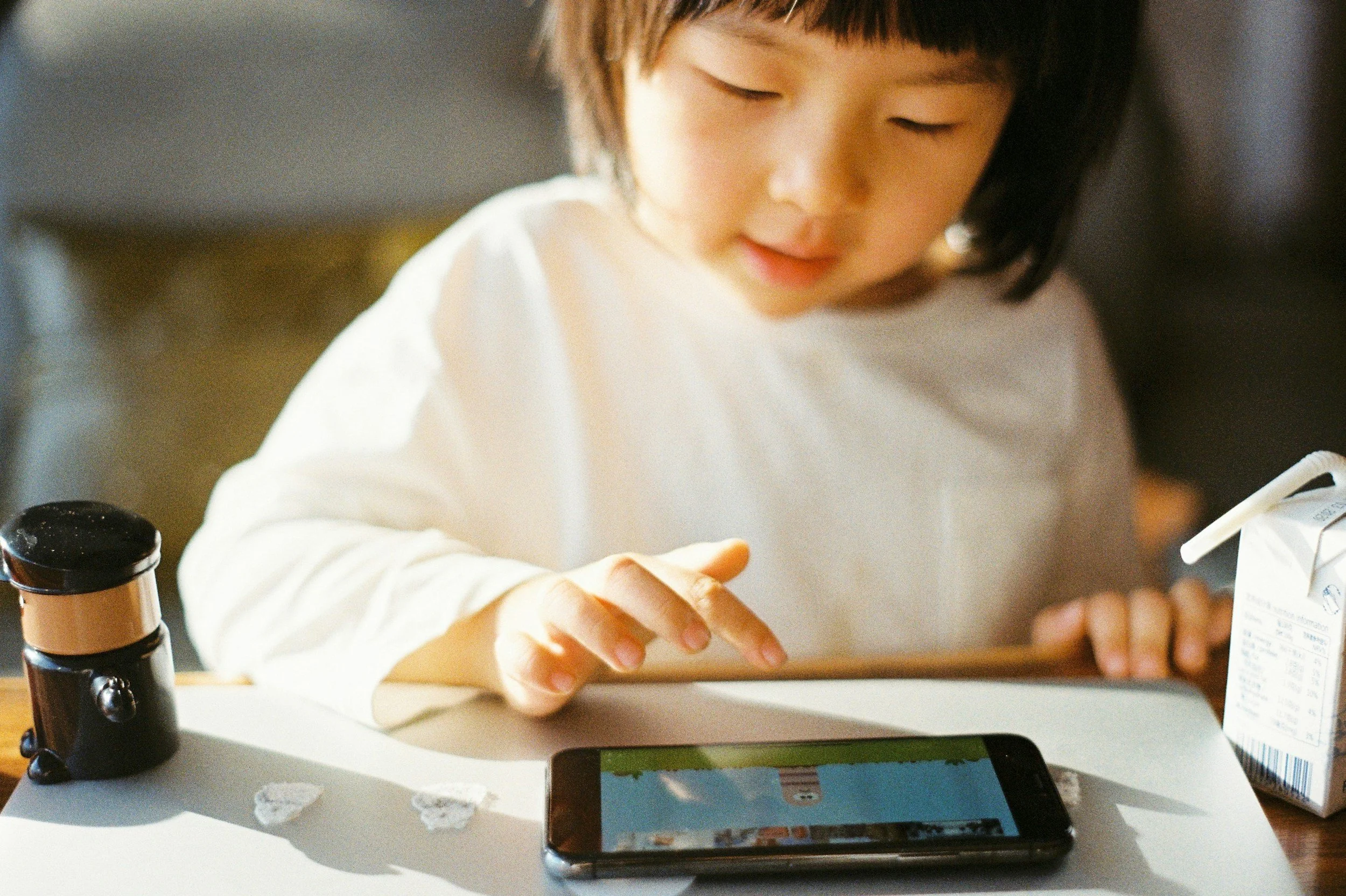 A young girl with black hair and a white shirt looks at her smartphone screen on a table, with a small black and gold telescope, crumpled paper, and a juice box nearby.