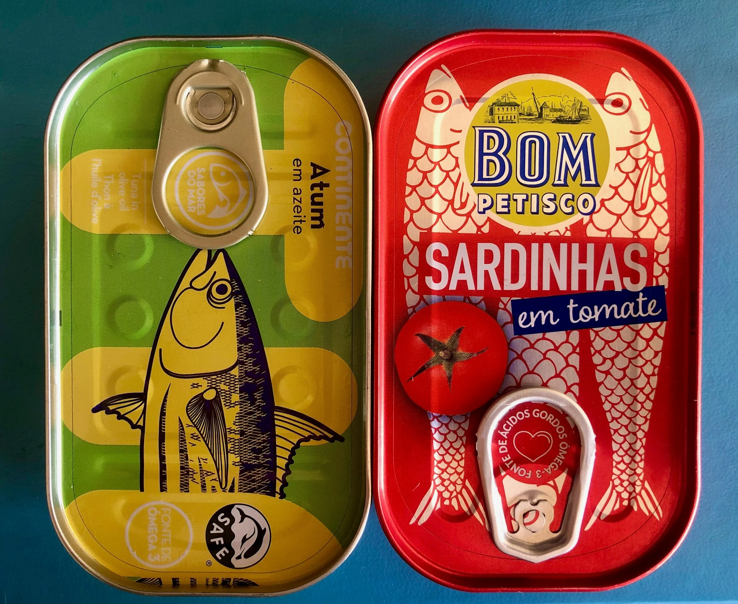 Canned sardines in tomato sauce on a red container with a tomato image, next to a yellow and green tin of sardines in sunflower oil with a fish illustration.