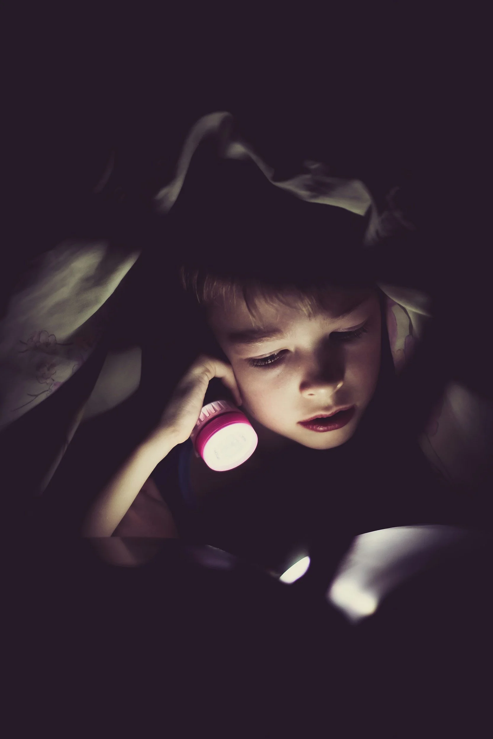 Child lying in dark with a flashlight, looking at a device with a curious expression.