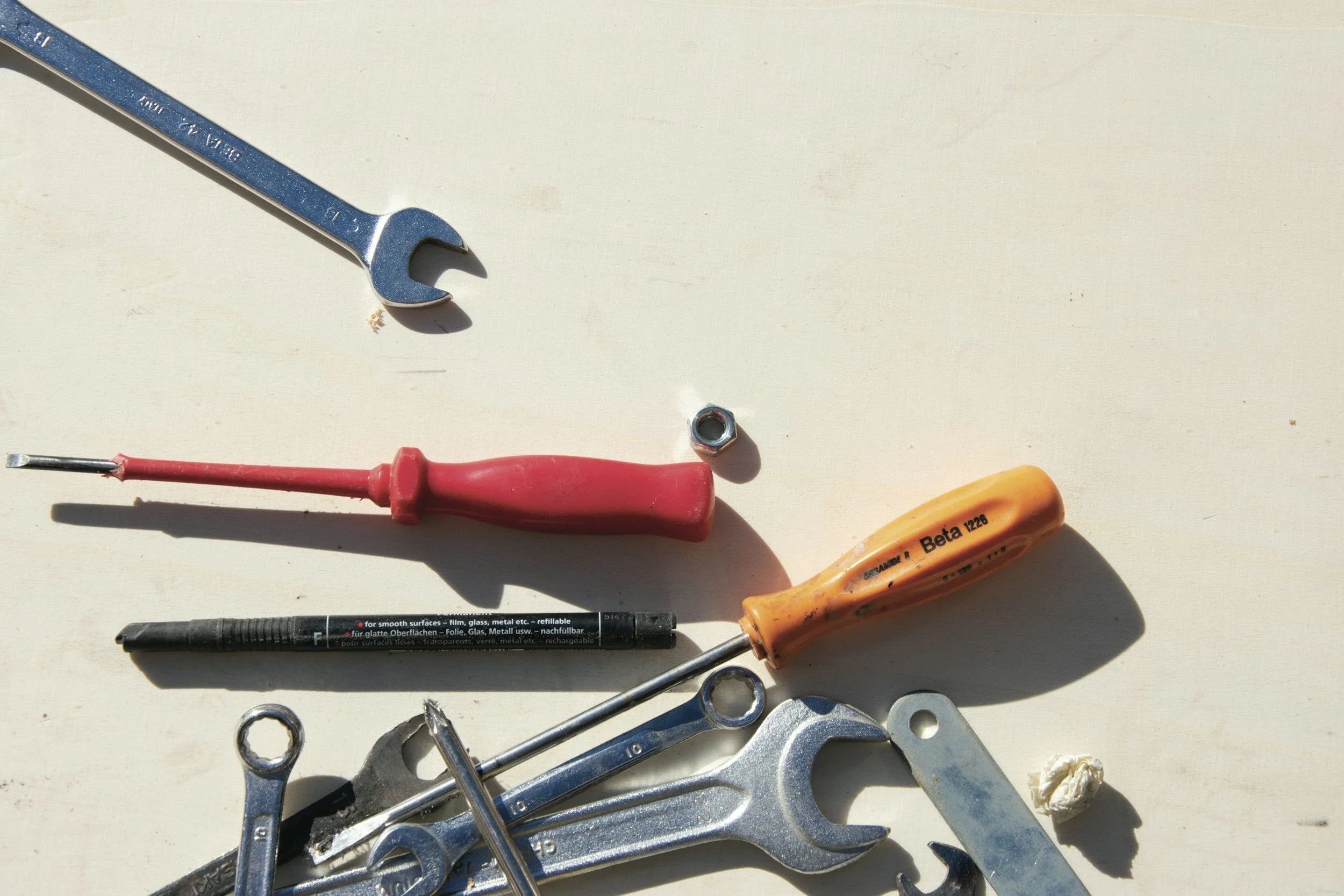 Assorted hand tools including wrenches, screwdrivers, a marker pen, a nut, and other small tools spread out on a beige surface.