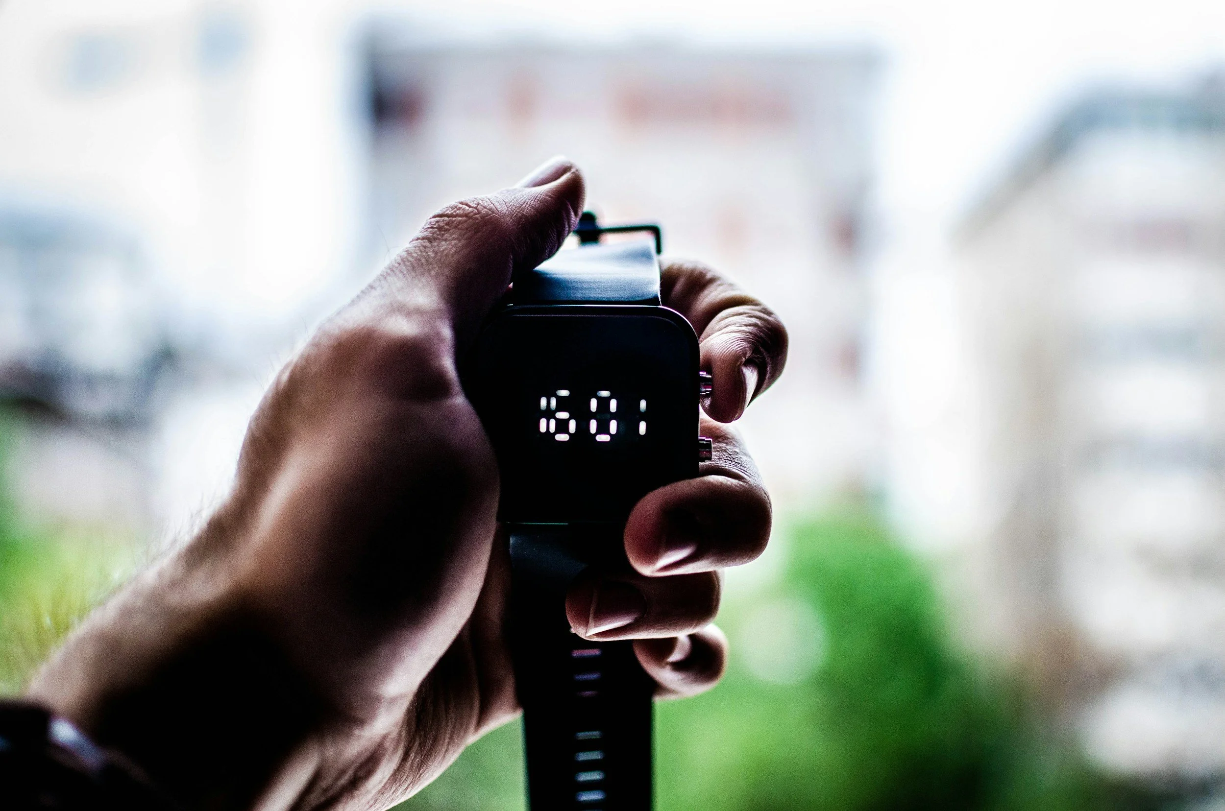 A person holding a digital wristwatch showing the time 15:01, with a blurred background of a window and outside environment.