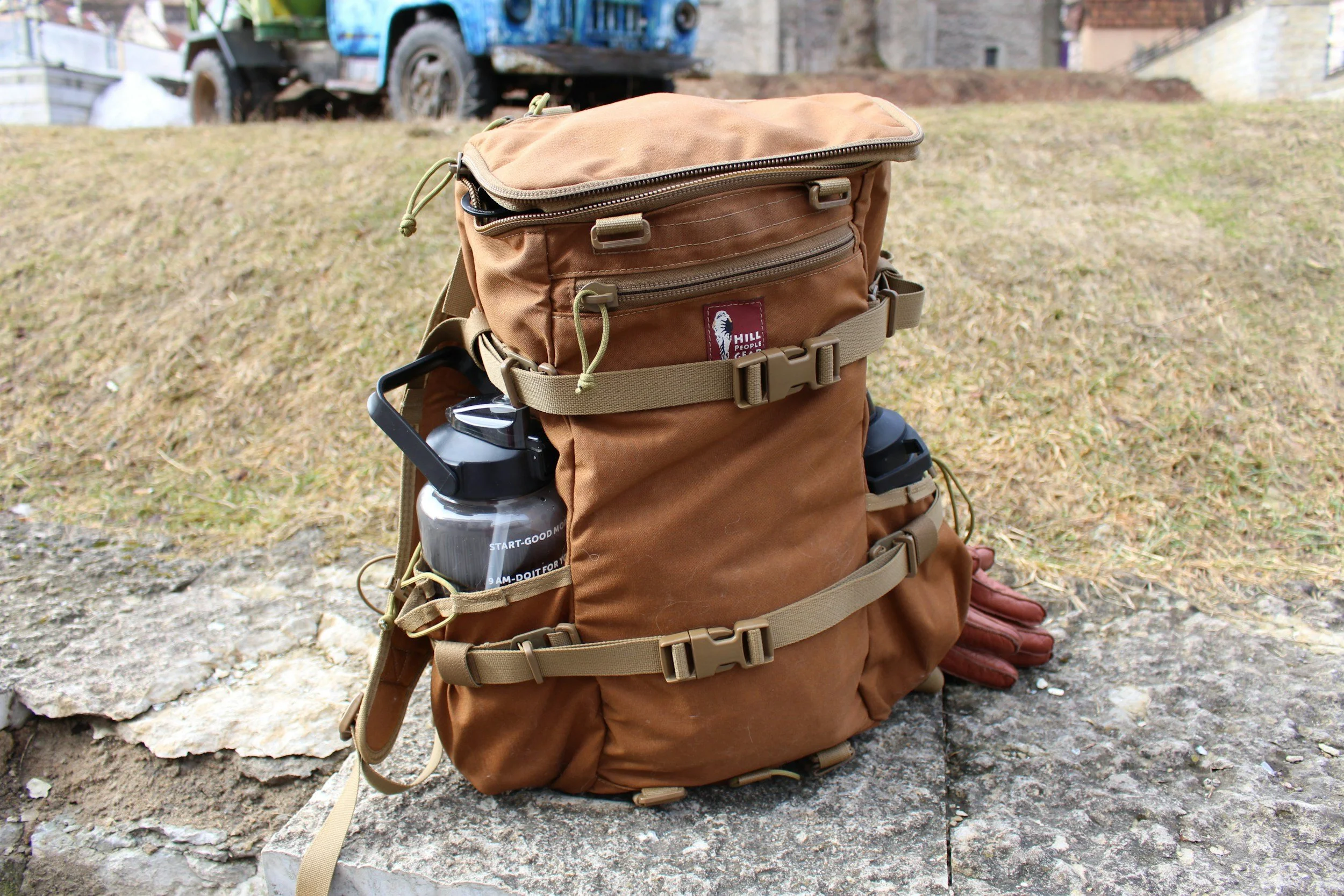 Brown hiking backpack with water bottle holder, placed on a stone surface outdoors with grass and a truck in the background.