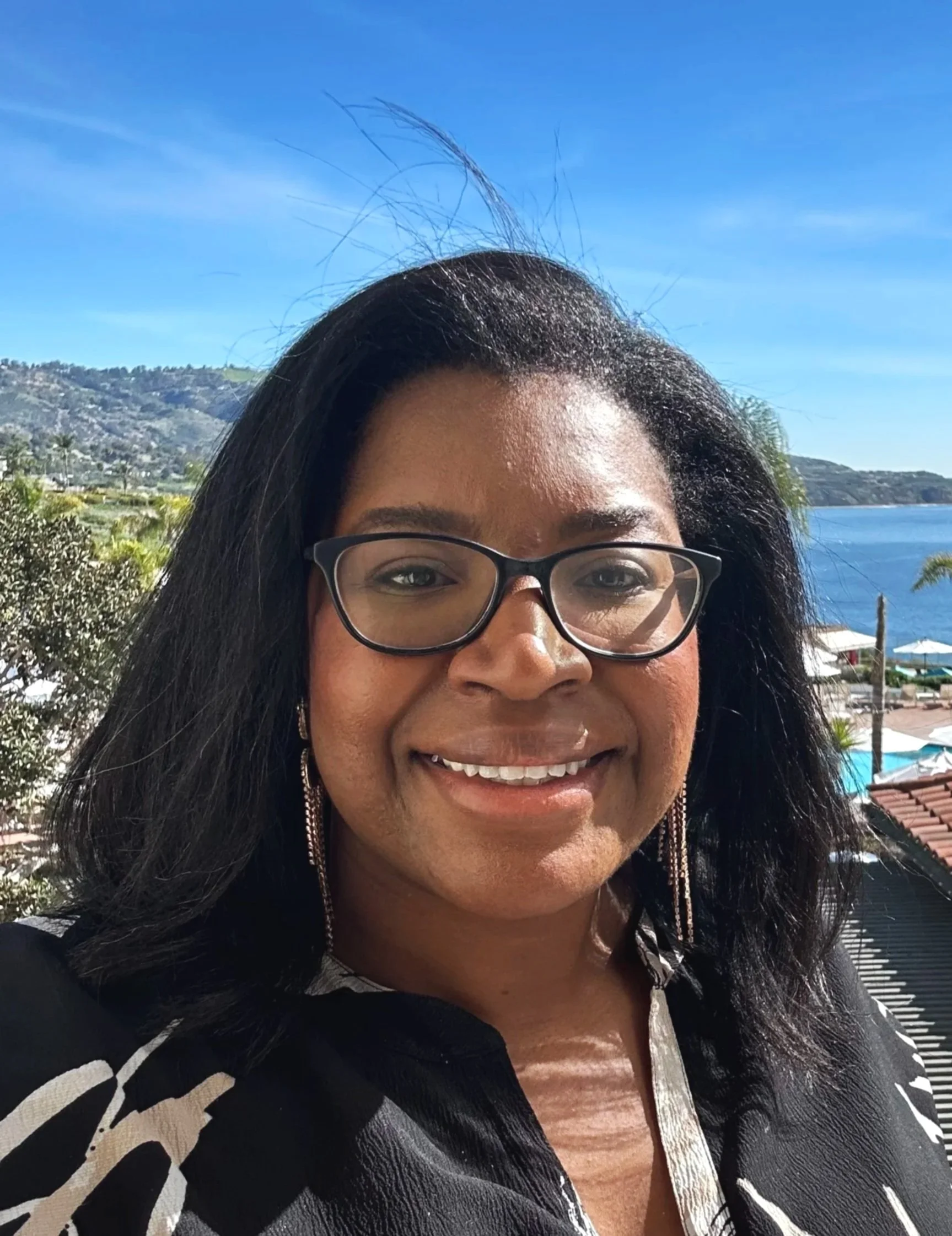A smiling woman with black hair and glasses standing outdoors with a view of the water and a hilly landscape in the background.
