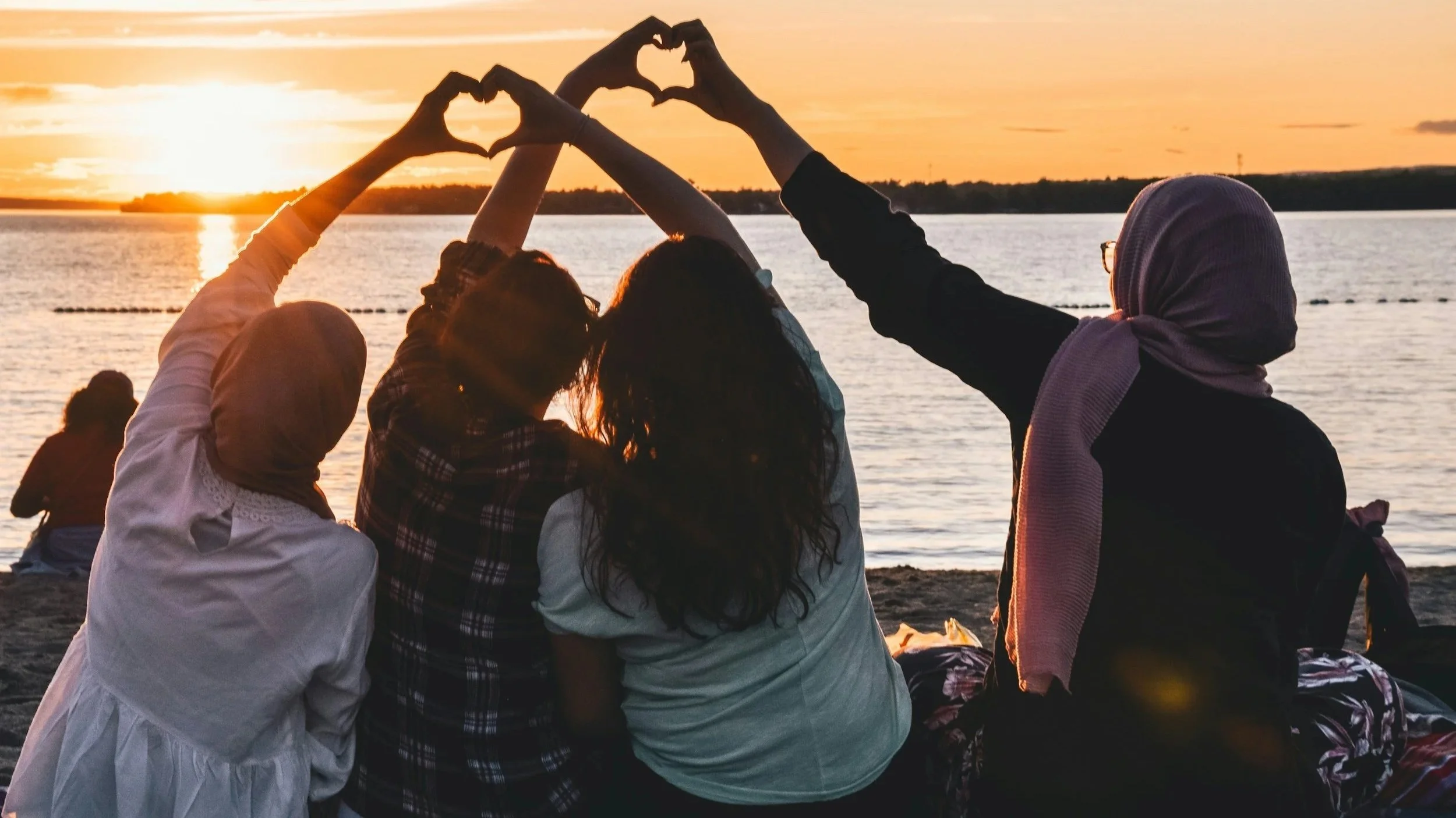 People sitting by a lake during sunset, making heart shapes with their hands
