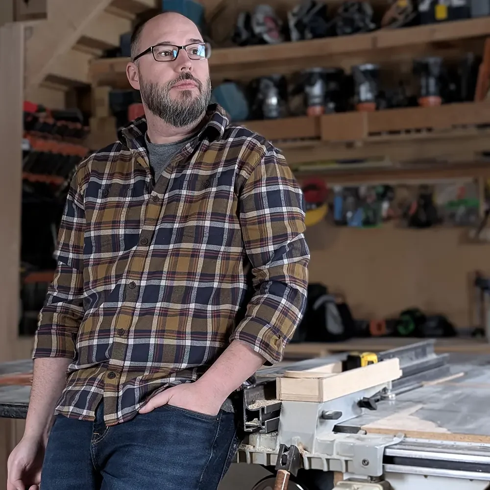 Jake Campbell (Owner of Campbell's Woodcraft) with glasses and a beard in a plaid shirt standing in a woodworking workshop, leaning against a table saw with his hand in his pocket.