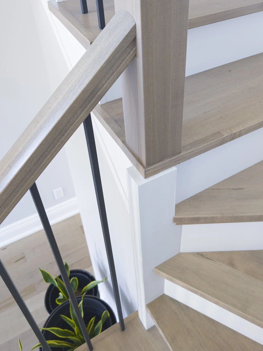 Looking down a winder staircase transformation featuring grey-toned hardwood recap treads, a matching custom handrail, modern matte black balusters, and crisp white risers. Potted plants provide contrast below.