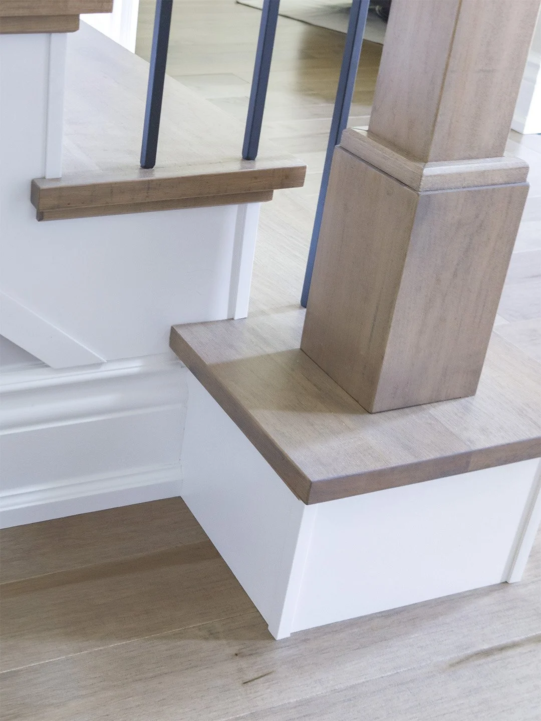 Close-up perspective of a custom-finished solid hardwood newel post matching the grey-toned recap treads. The post integrates perfectly with the built-up starting tread block, alongside modern matte black square metal balusters and clean white painte