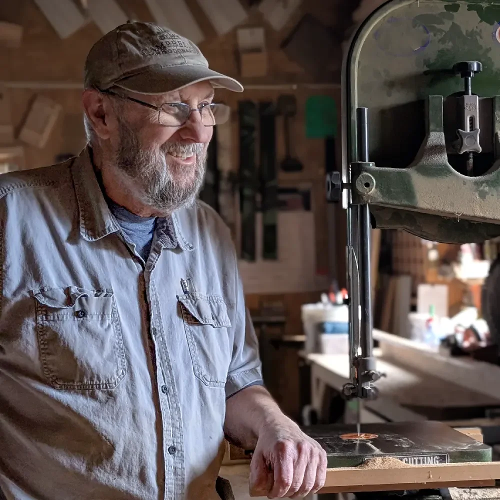 Marty Campbell (Owner of Campbell's Woodcraft) with a beard and glasses, wearing a cap and a light gray button-up shirt, is smiling as he works with a bandsaw in a woodworking shop.