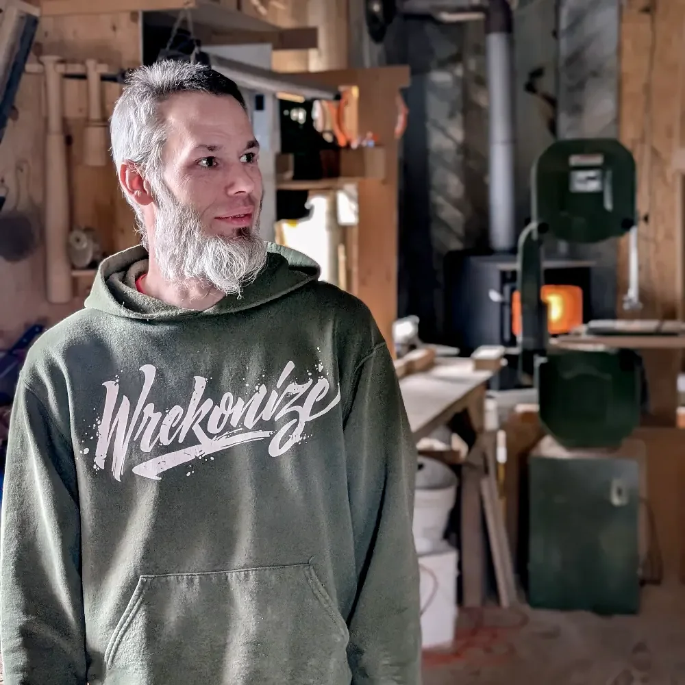 Jay Campbell with gray hair and beard wearing a green hoodie standing inside a wood workshop with various tools and equipment in the background.