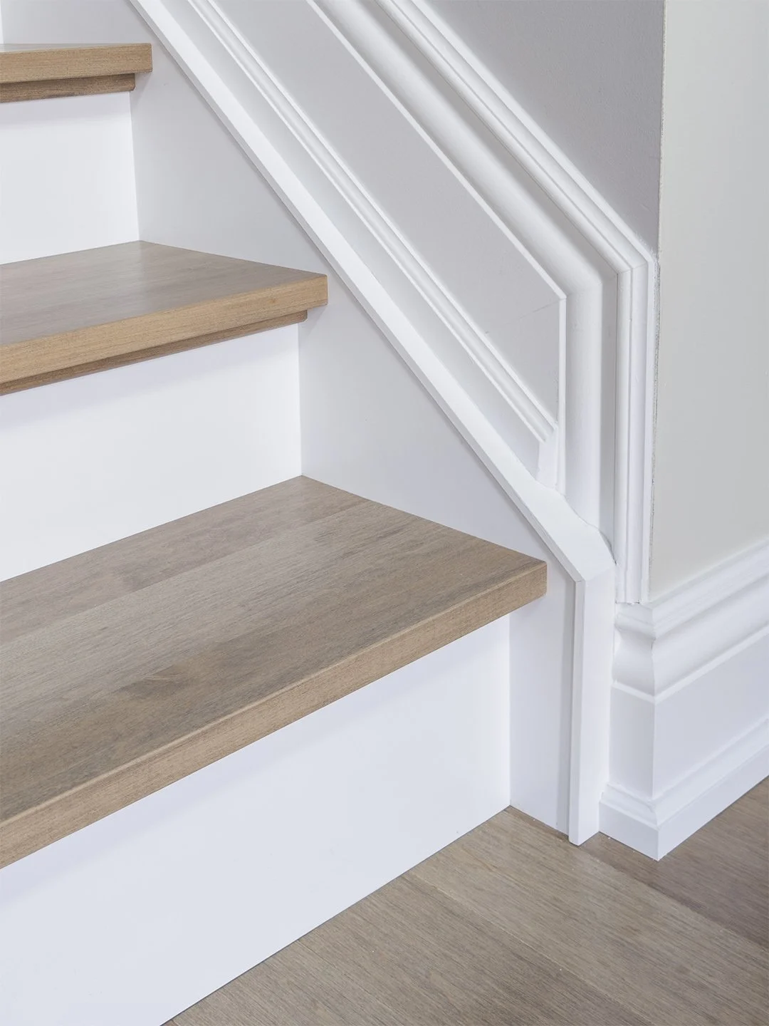 Detailed photograph capturing the immaculate junction between a grey-toned hardwood recap tread, crisp white risers, and intricate traditional multi-profile baseboard and skirt board trim, showing seamless craftsmanship.