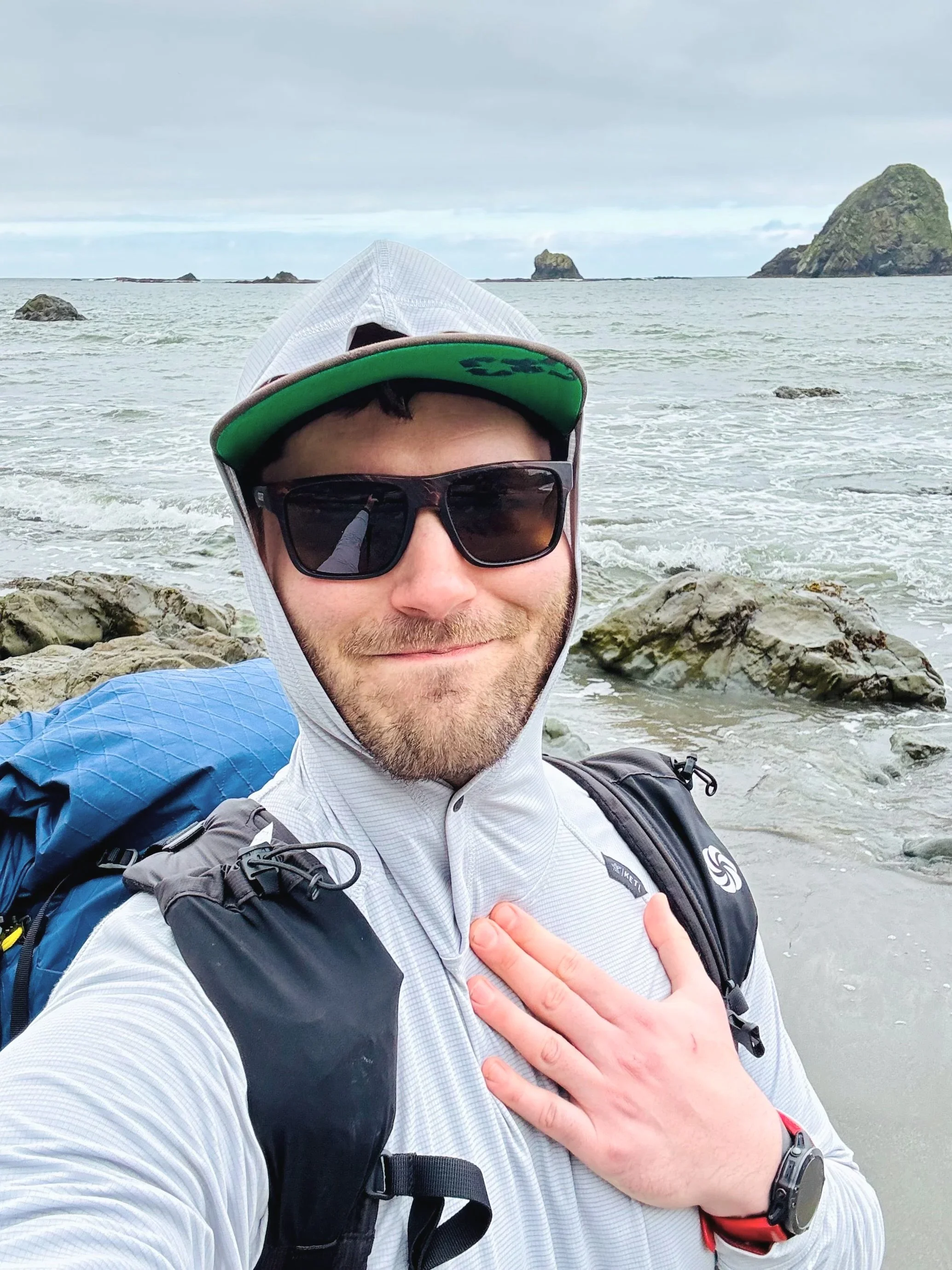 A man taking a selfie on a rocky beach with the ocean and small islands in the background, wearing sunglasses, a cap, a light jacket, and a backpack.