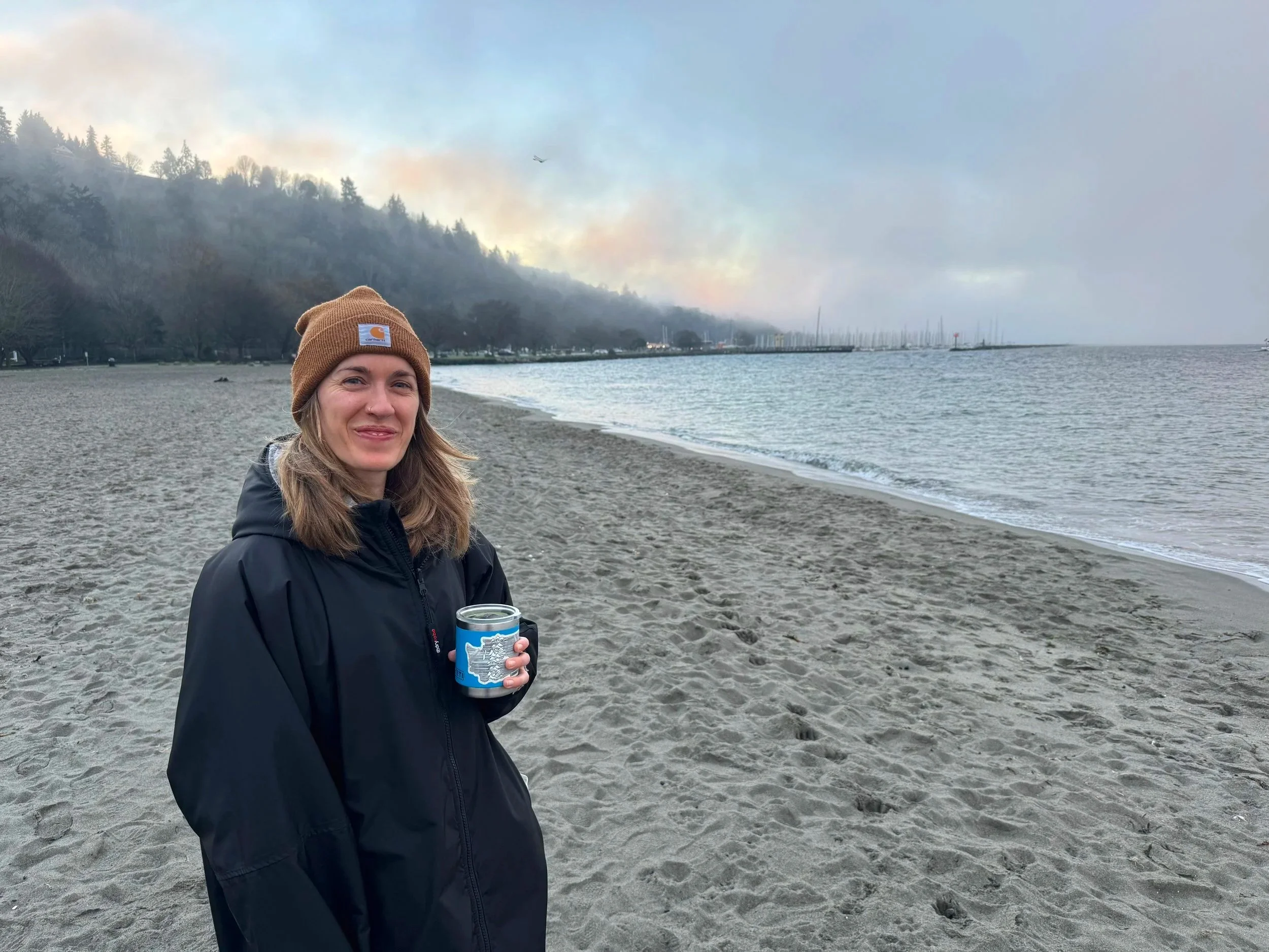 A woman in a brown beanie and black jacket standing on a beach with water and a forested hillside in the background. She is holding a can and smiling, with a misty sky overhead.
