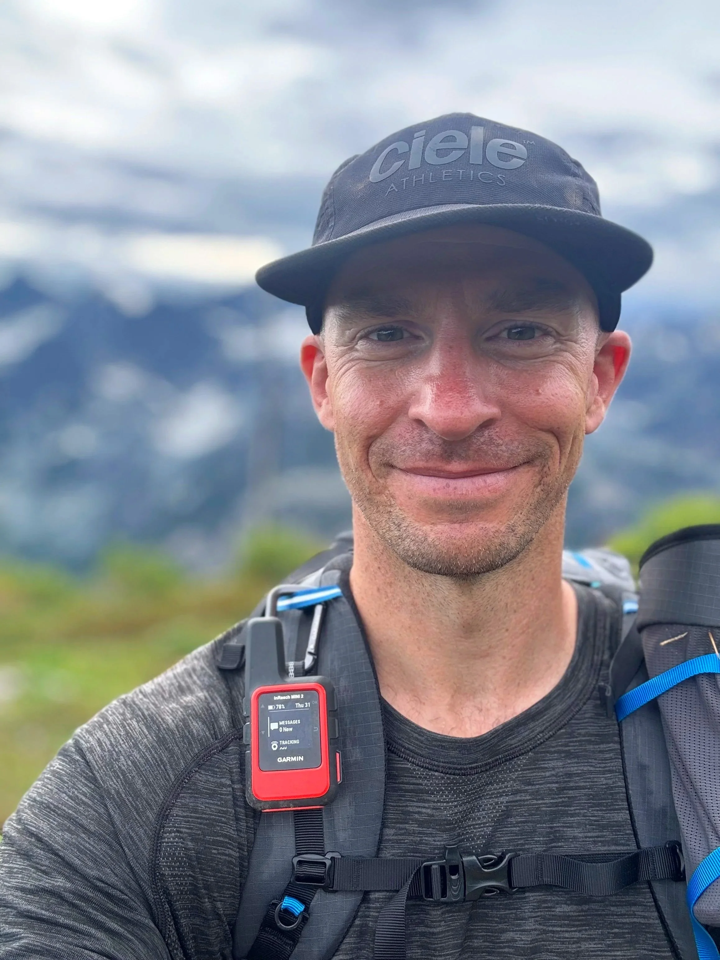 Close-up of a smiling man wearing a black cap, carrying a backpack with a Garmin GPS device attached, set against a background of cloudy sky and mountains.