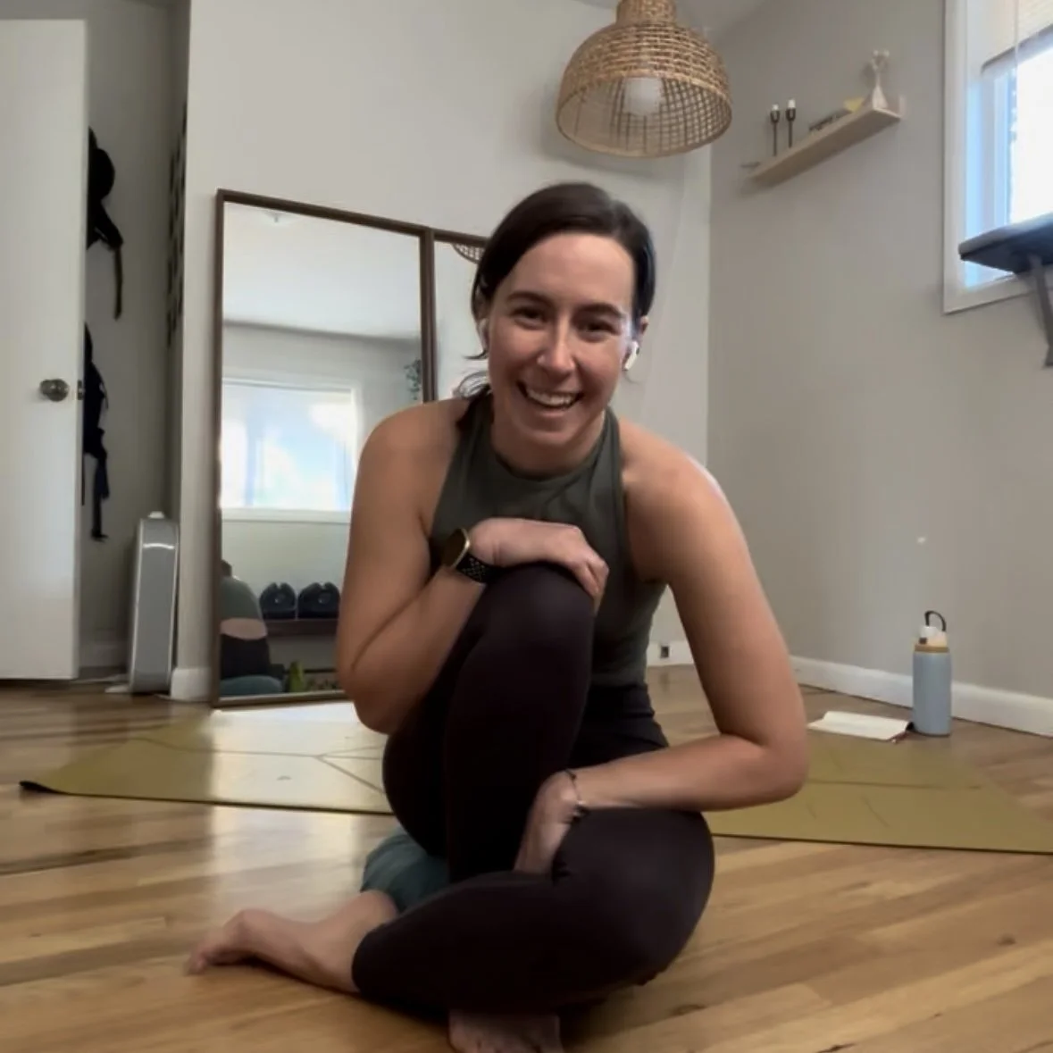 A woman practicing yoga in a home studio, smiling and sitting on a yellow yoga mat with one leg crossed over another, in front of a mirror and decorative items.