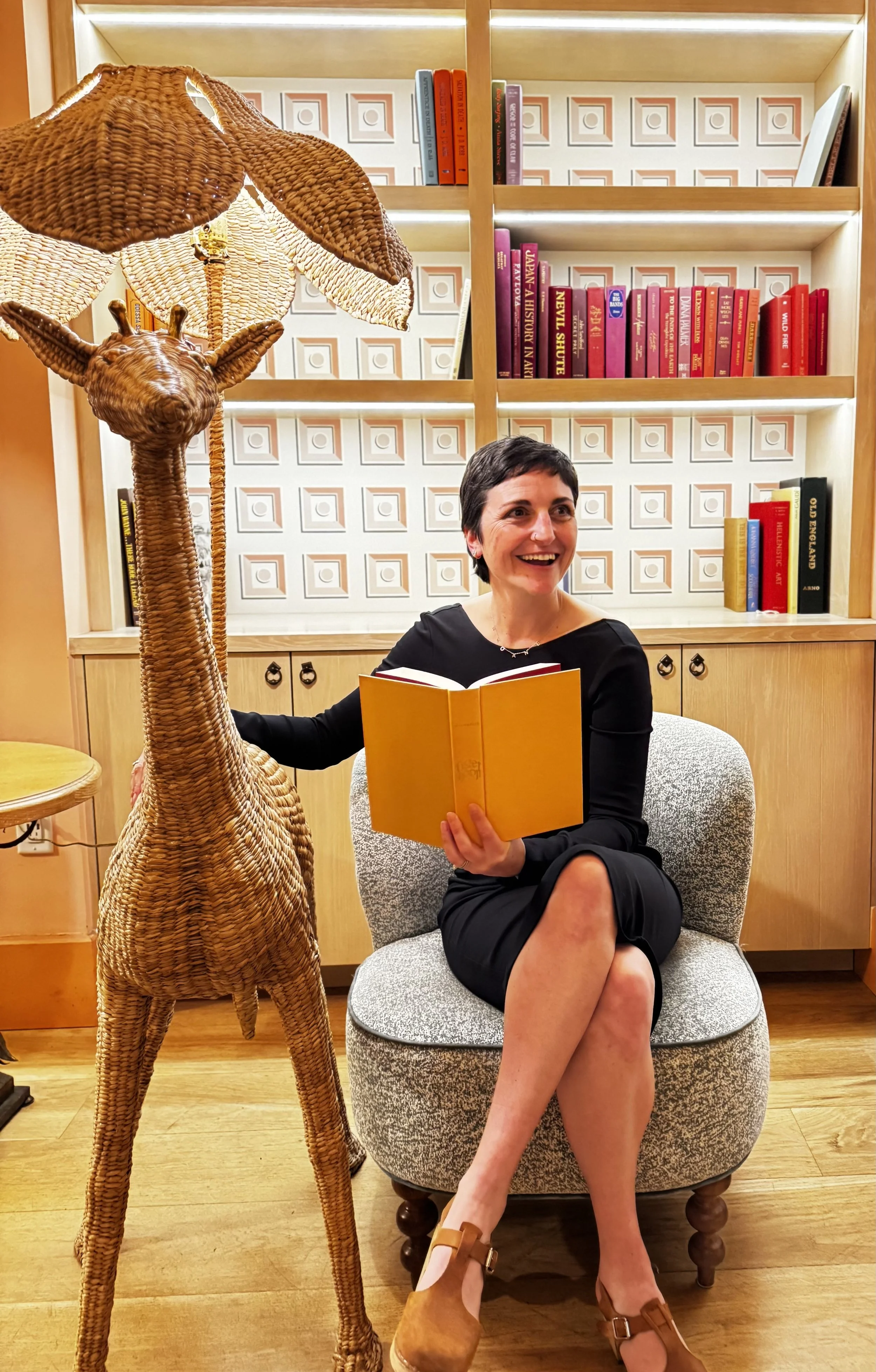 A woman with short dark hair and a black dress sitting on a gray armchair in a room with wooden floors and bookshelves, holding an open yellow book and smiling. Next to her is a tall wicker giraffe sculpture. Behind them are shelves filled with colorful books, and the room has warm lighting.