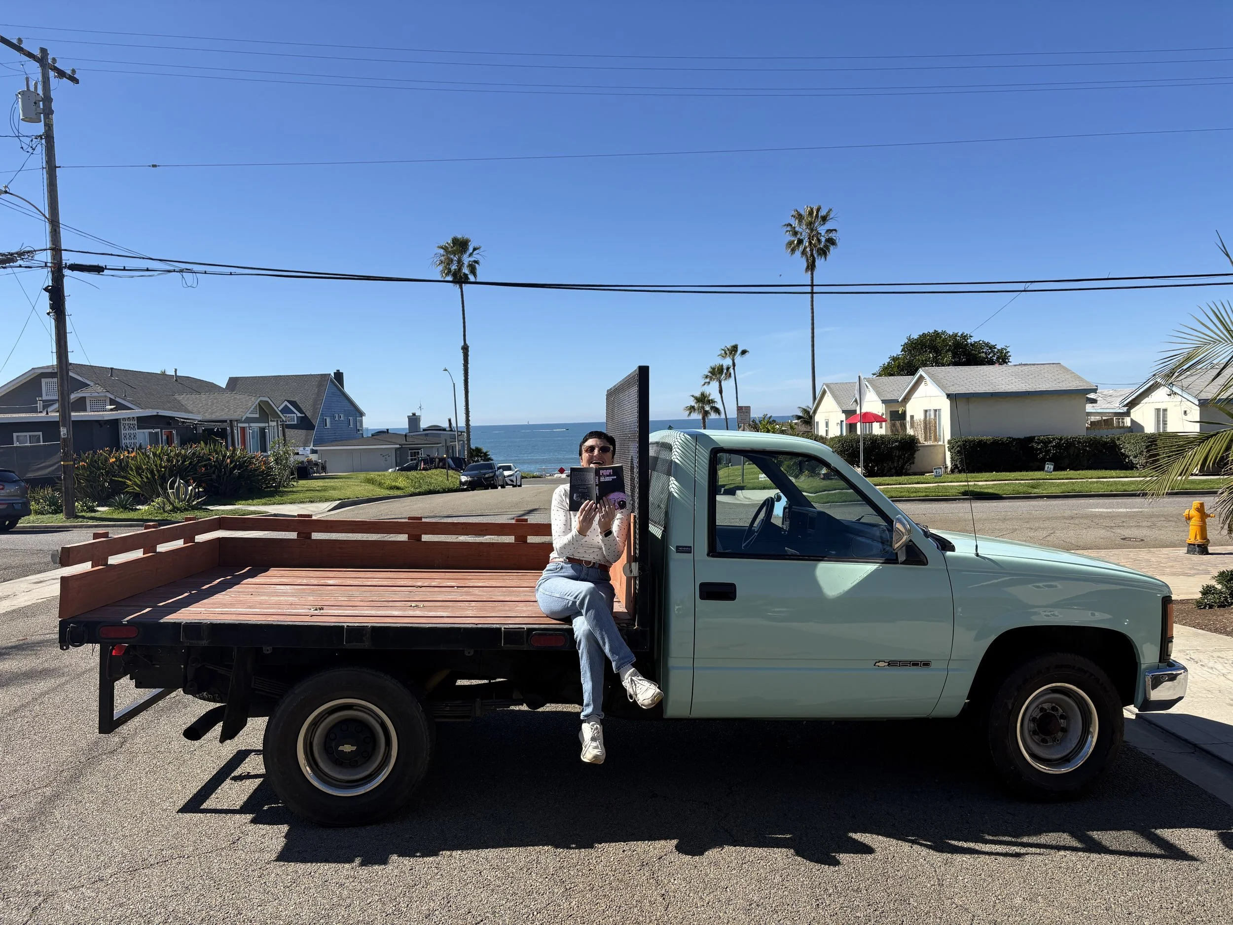 A woman sitting on the edge of a light green pickup truck's cargo bed, reading a book, with the ocean and palm trees in the background under a clear blue sky.
