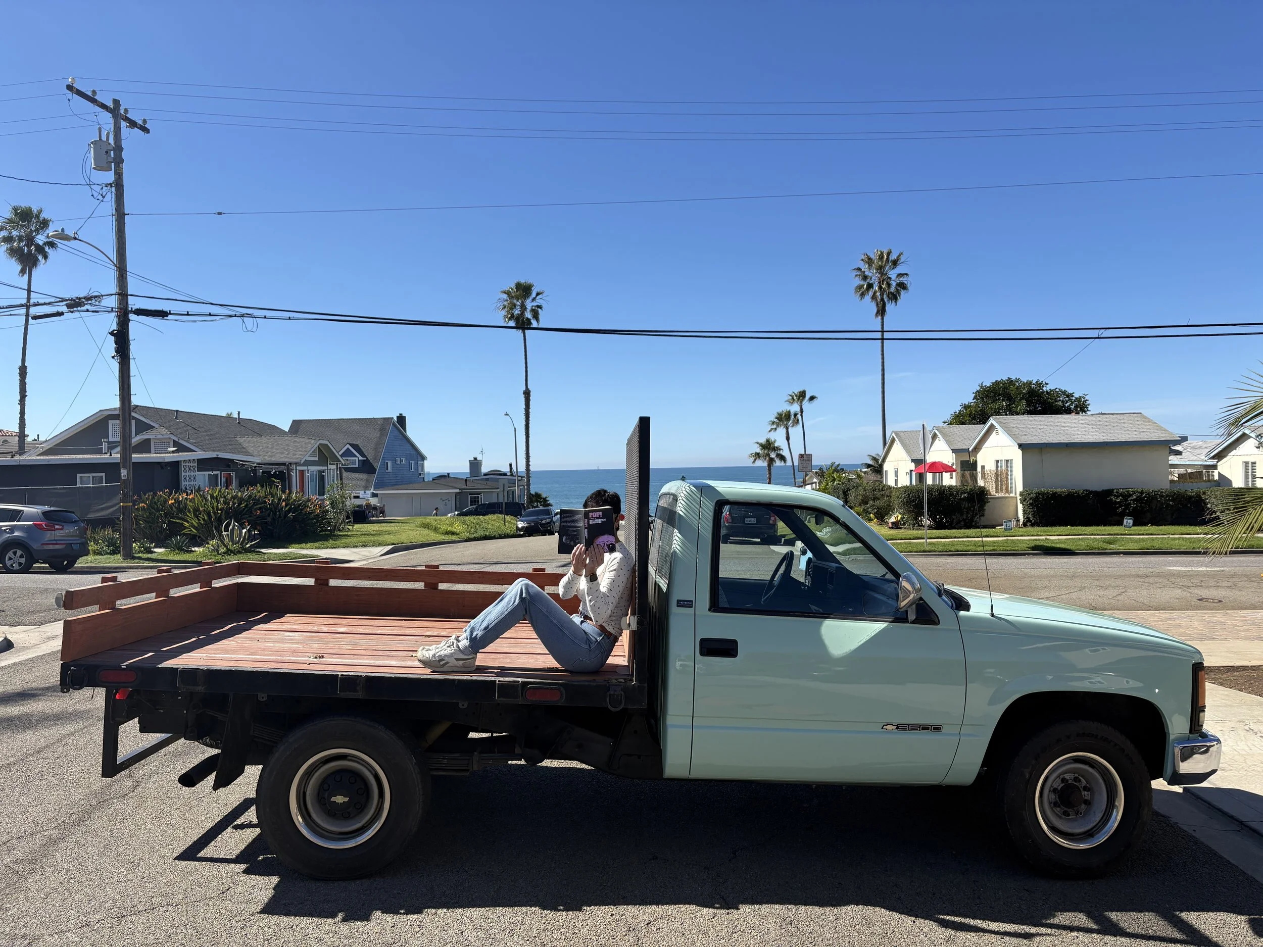 A woman in jeans and a white sweater sitting in the back of a light blue pickup truck, reading a book on a sunny day with clear blue skies, palm trees, and houses in the background.