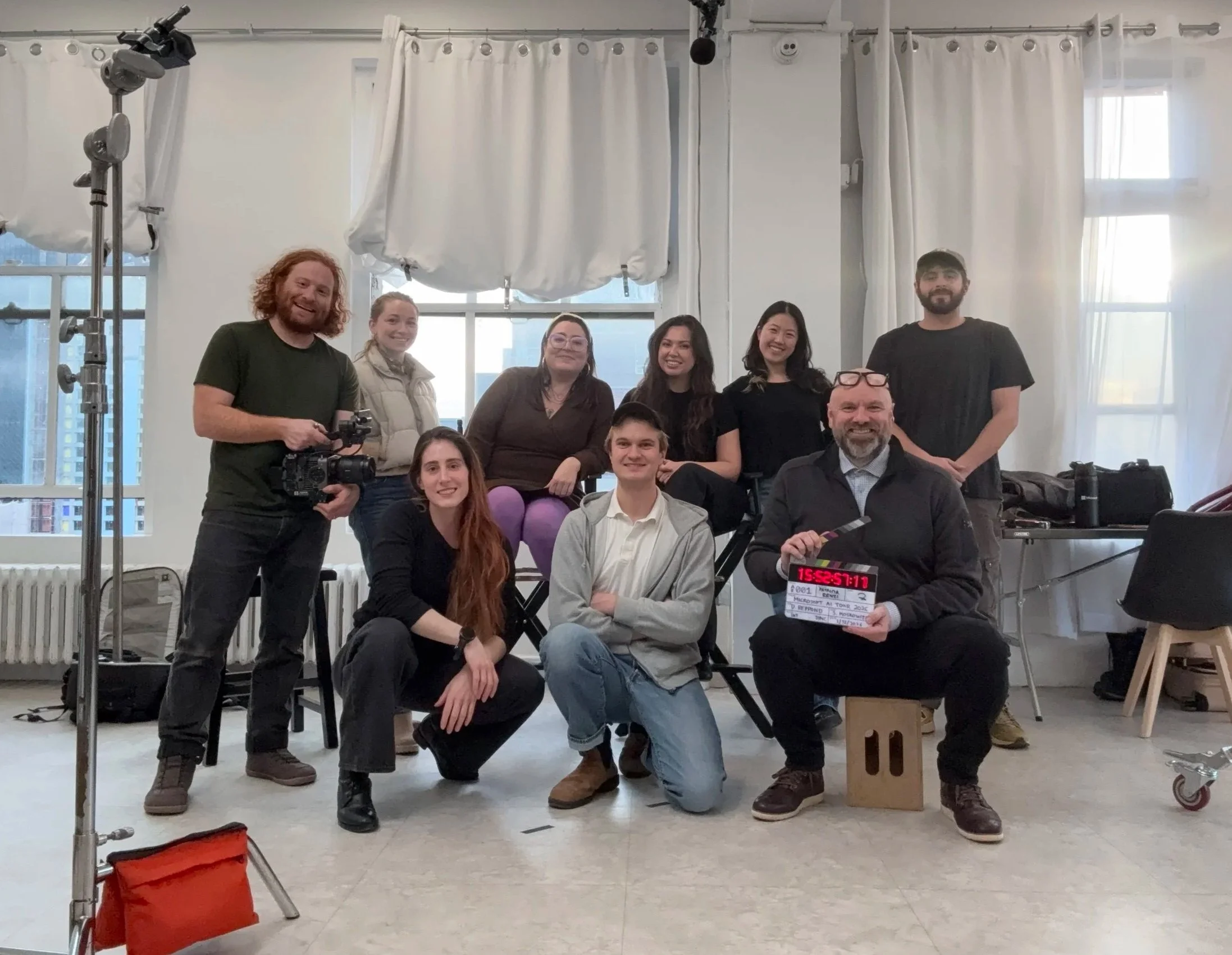 Group of ten people, including a man with a clapperboard, posing in a bright room with white curtains and large windows, possibly on a film or video set.