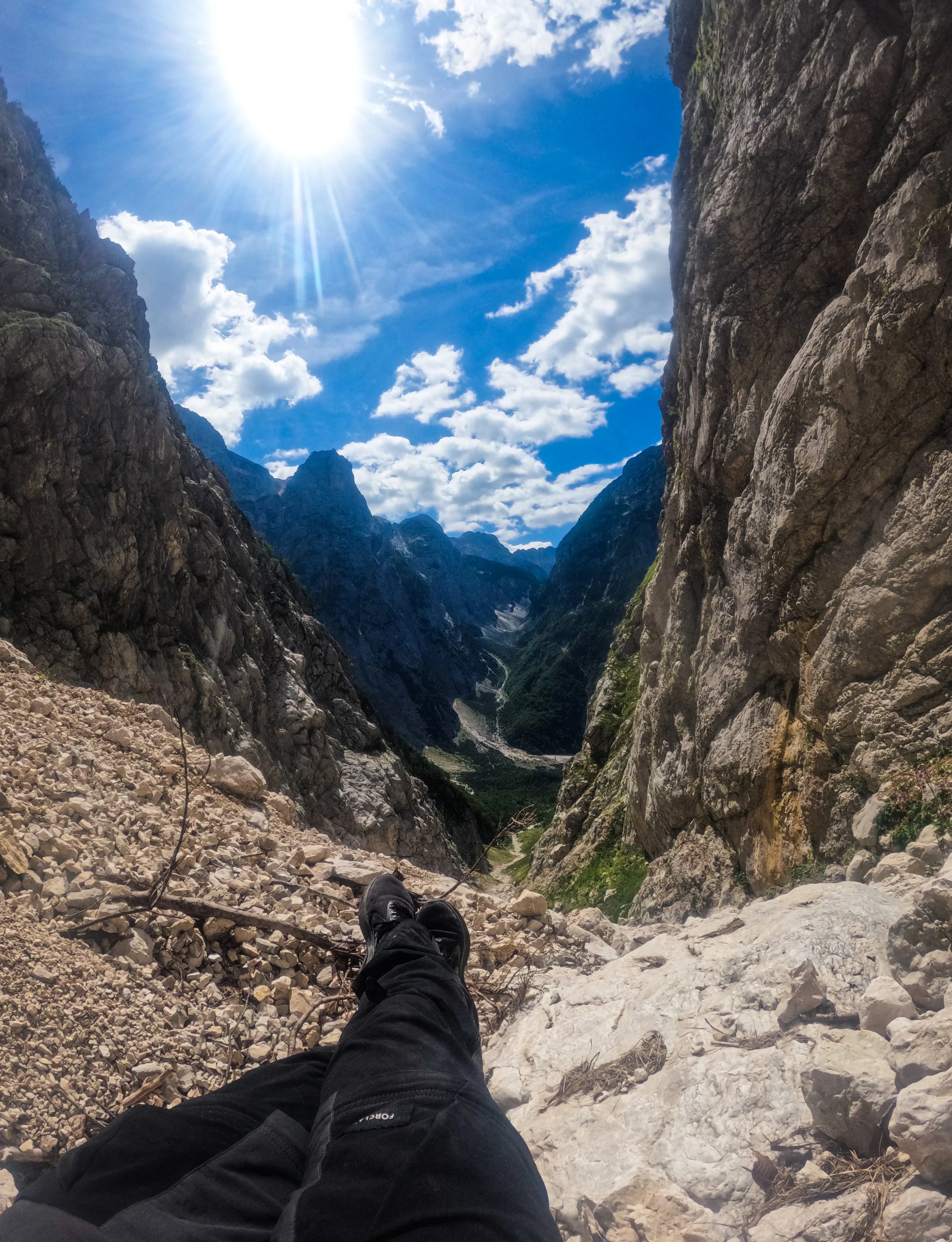 Person resting on a rocky mountain slope, overlooking a deep valley with steep cliffs and a river below, under a bright sun and partly cloudy sky.