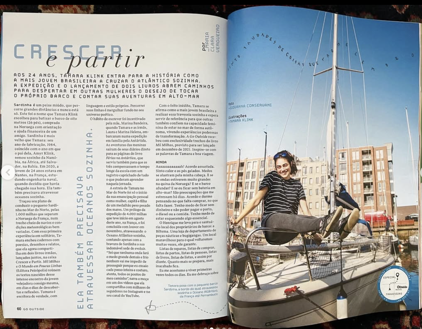 Sailor sitting on the deck of a sailboat moored in a marina with modern buildings in the background, wearing a striped shirt and leggings, smiling at the camera.