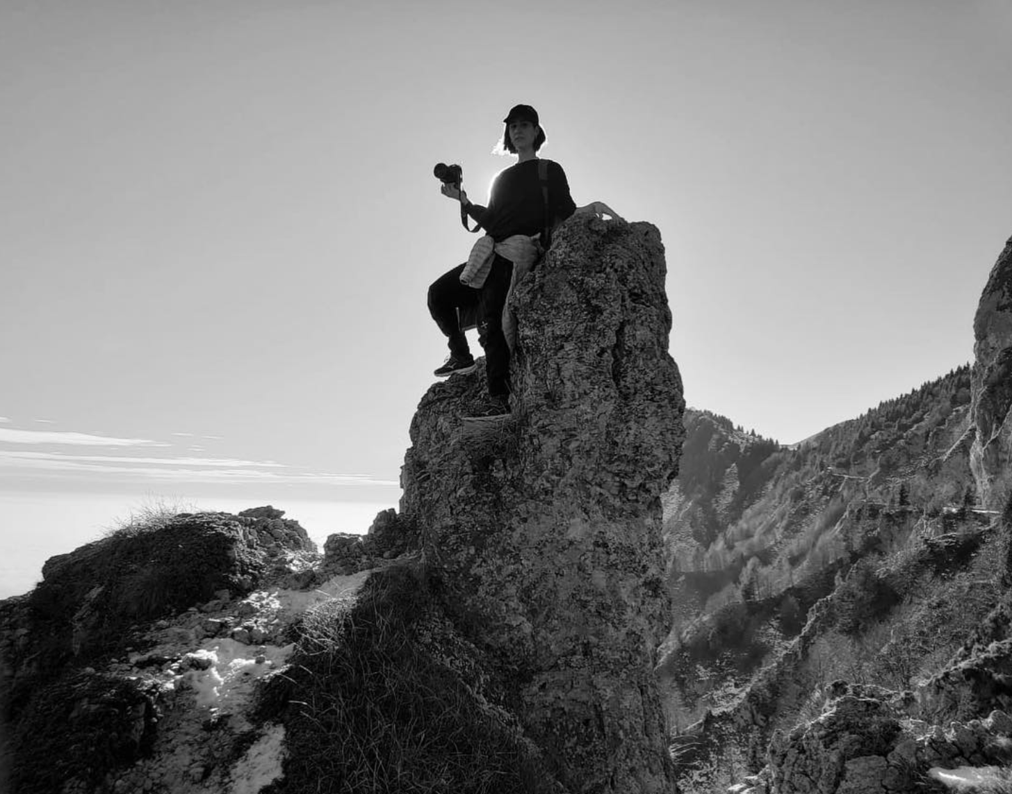 Person sitting on top of a large rock formation in a mountain landscape, holding a camera, with mountains and a partly cloudy sky in the background.