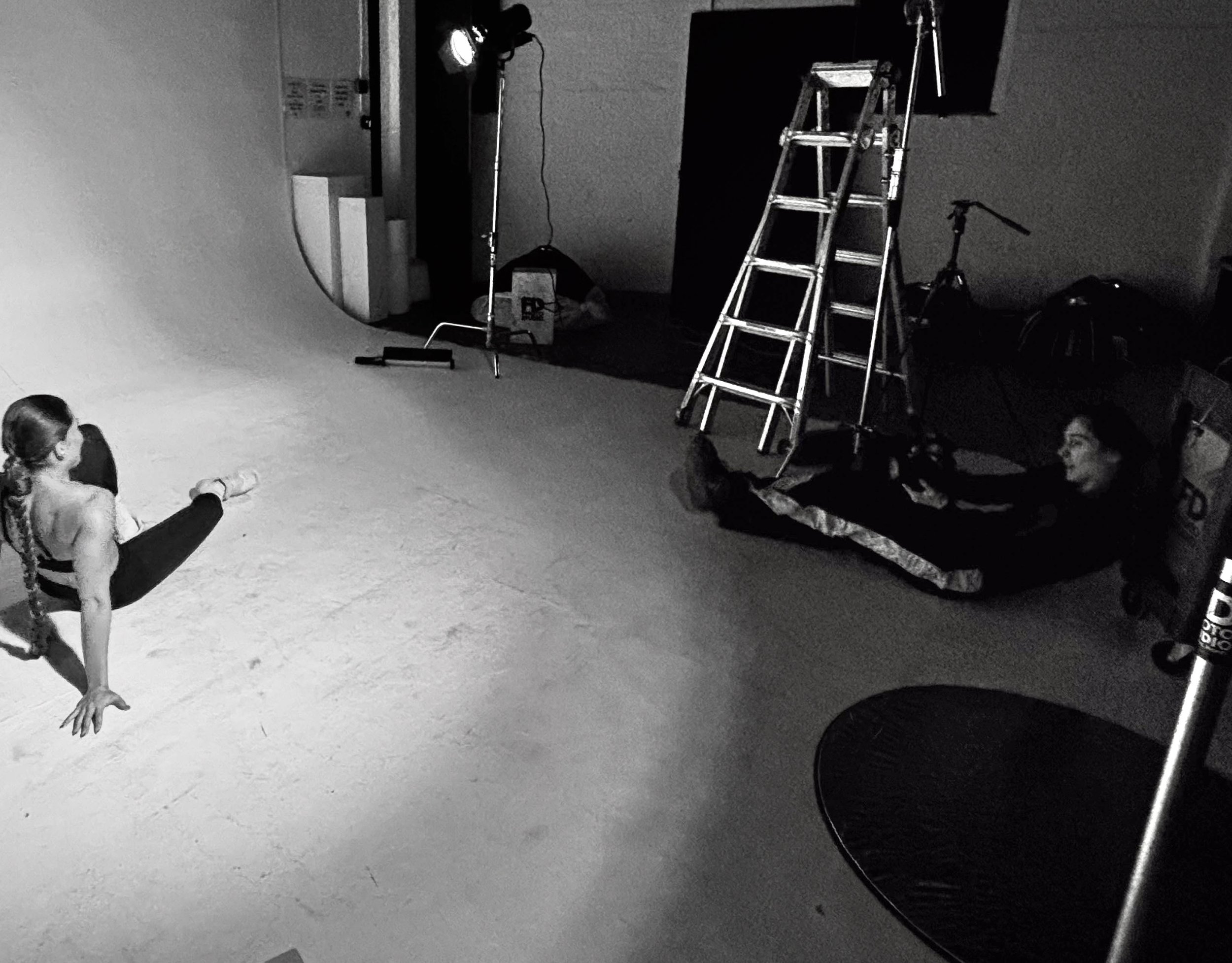 A woman in a photography studio with a white backdrop, while a woman sit on the floor near by is taking pictures.