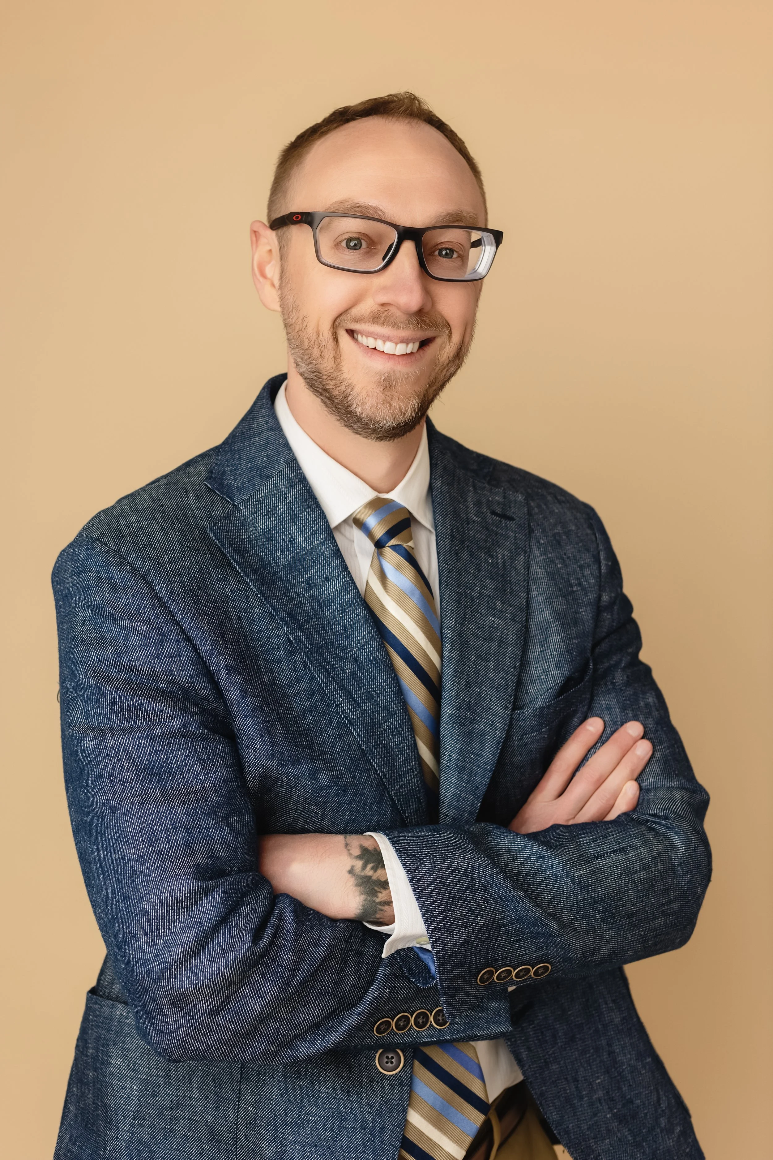 A man with glasses smiling, dressed in a blue suit with a striped tie, standing against a beige background with arms crossed.