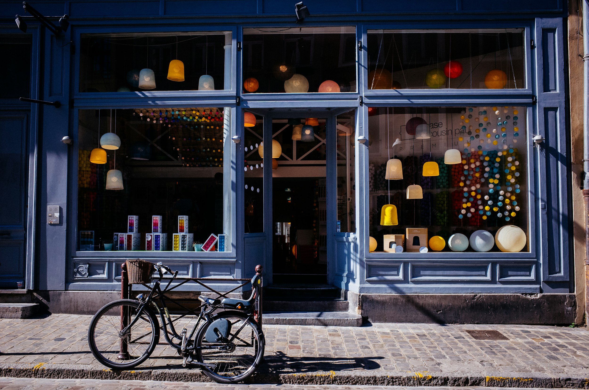 A storefront with large glass windows displaying various colorful lighting fixtures and decorative objects inside, with a bicycle parked on the cobblestone sidewalk in front.