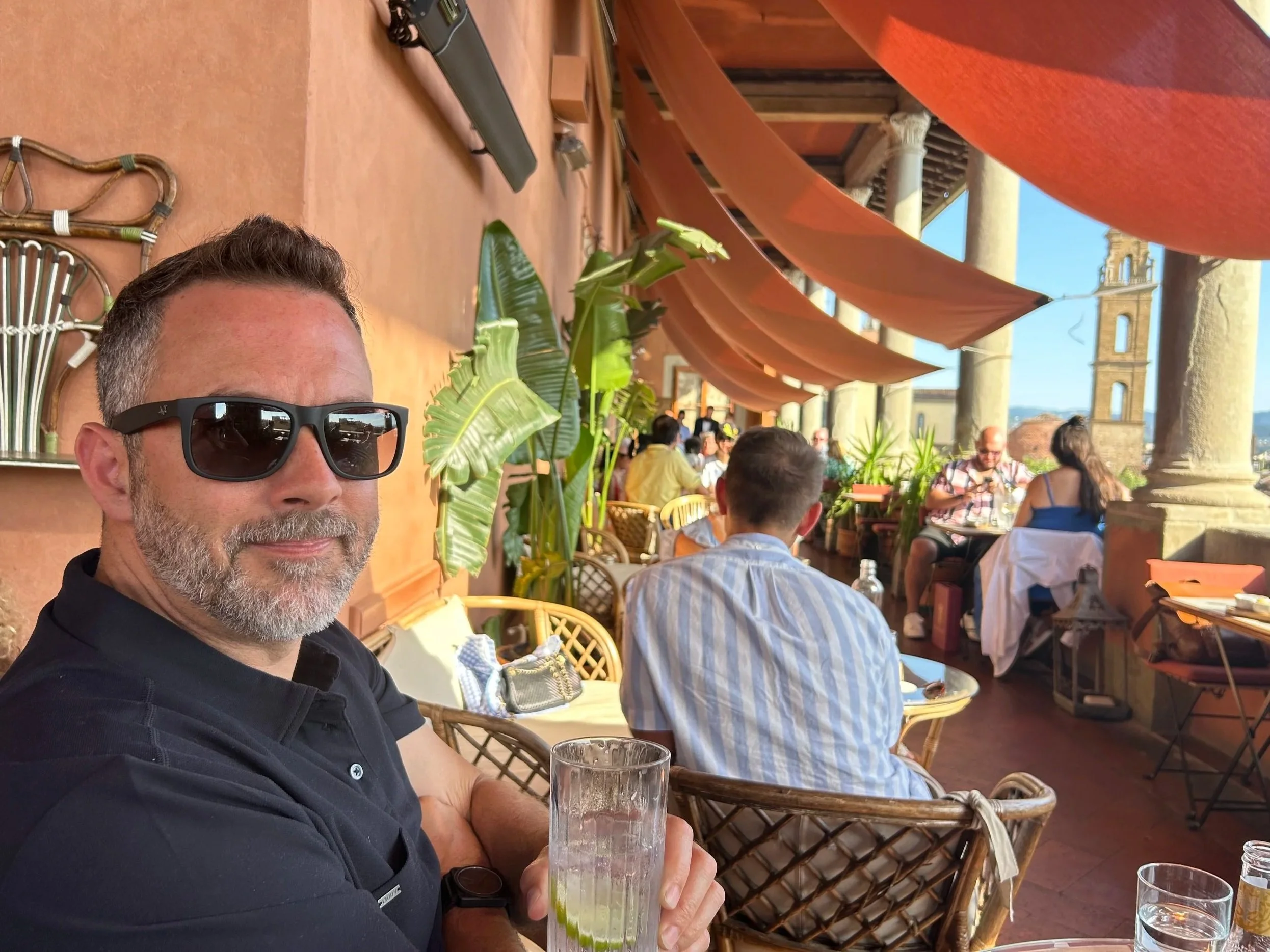 A man with short hair and a beard, wearing sunglasses and a black polo shirt, sitting at an outdoor restaurant with a glass of lime-infused water, with other diners and terracotta-colored awnings in the background, overlooking historical arches.