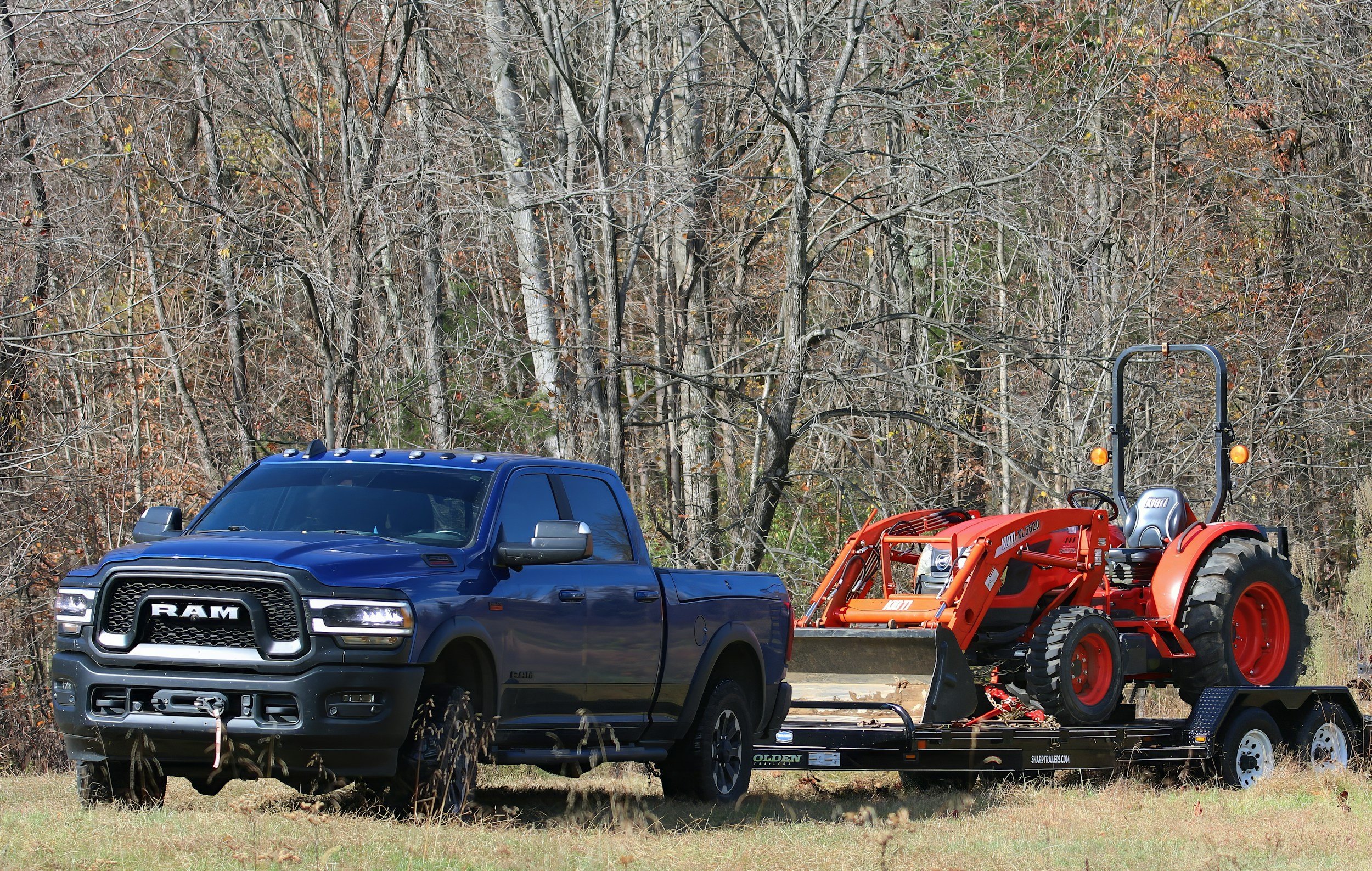 A blue RAM pickup truck on a trailer carrying an orange tractor in a grassy field with trees in the background.