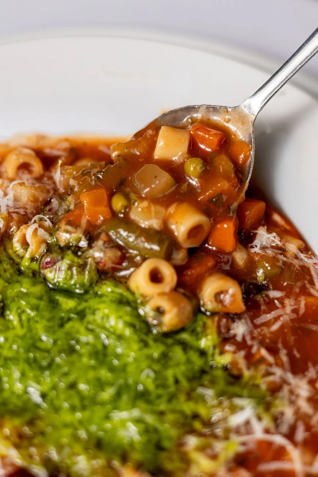 Close-up of a spoonful of vegetable soup with pasta, peas, carrots, and herbs, being lifted from a bowl with broccoli and shredded cheese visible in the background.