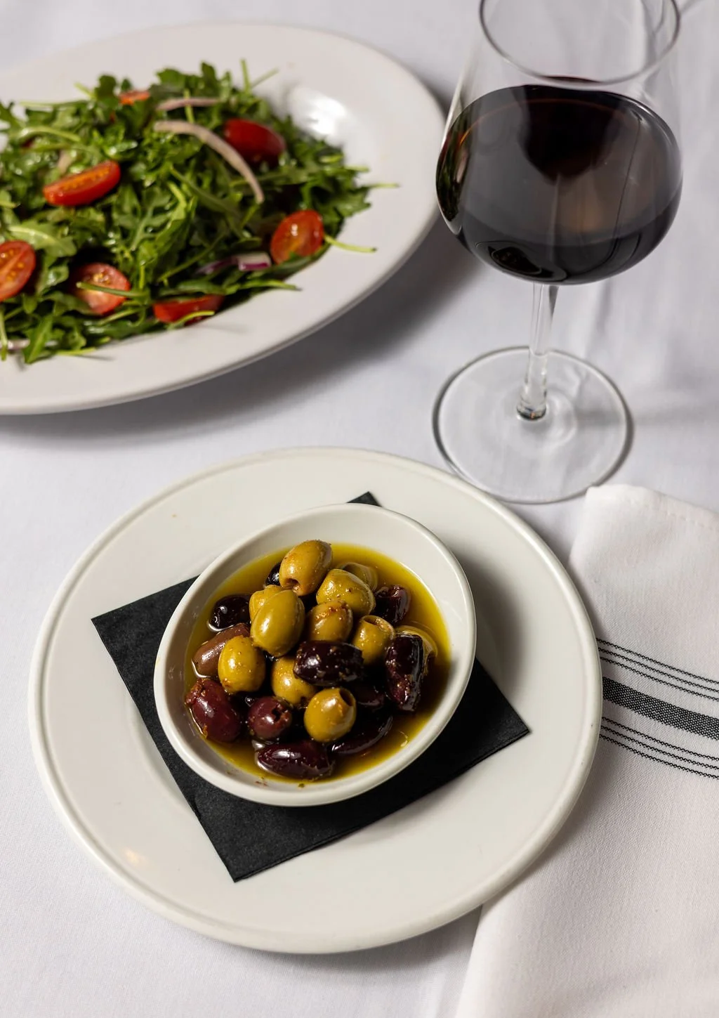 A table with a glass of red wine, a green salad with cherry tomatoes, and a bowl of green and black olives in oil.