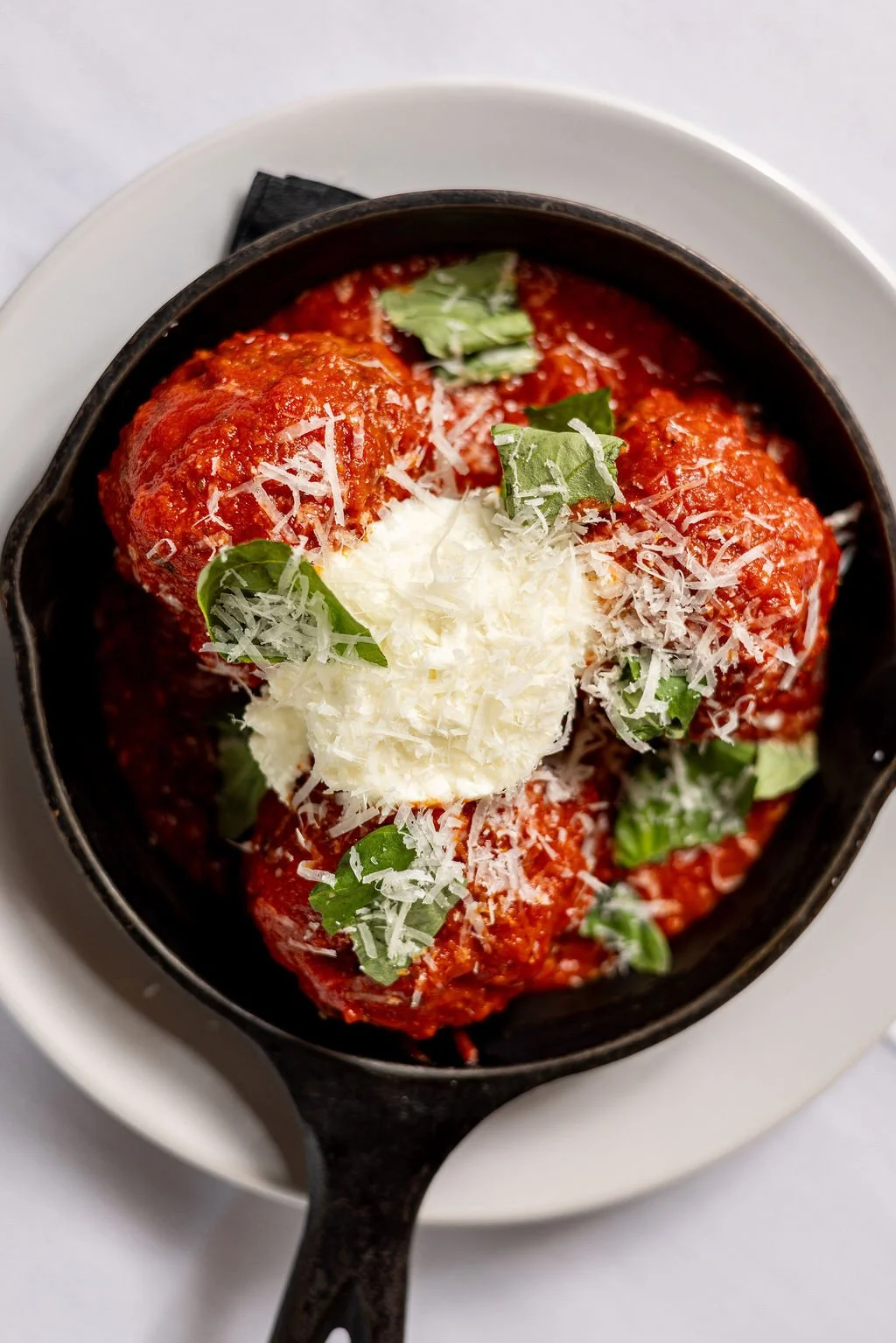 Three meatballs covered in marinara sauce, topped with grated cheese and fresh basil, in a black cast iron skillet.