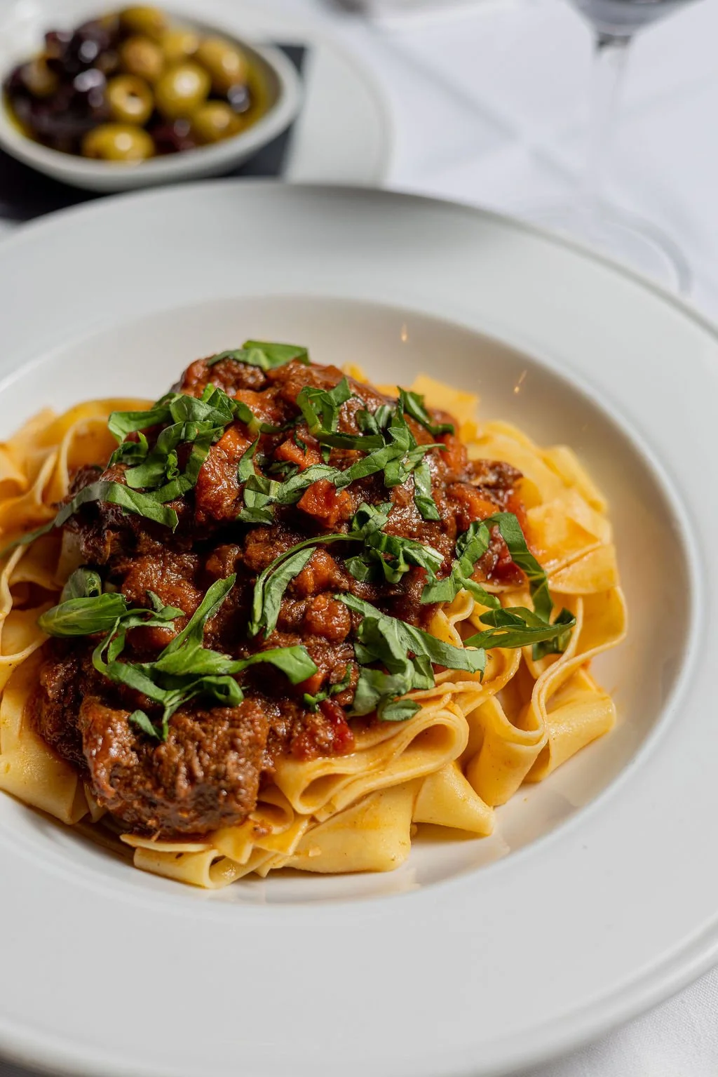 Plate of fettuccine pasta topped with meat sauce and chopped basil, with a bowl of mixed olives in the background.