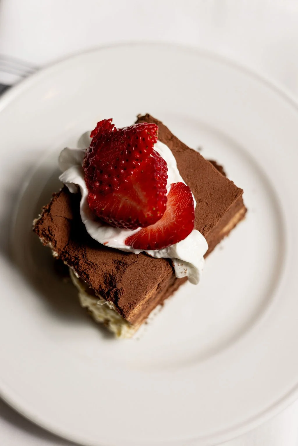 Square piece of chocolate cake topped with whipped cream and sliced strawberries on a white plate.