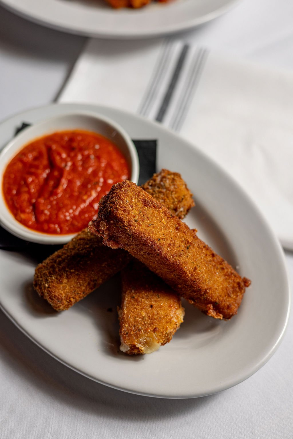 Plate with three fried mozzarella sticks and a small bowl of marinara sauce, on a white table.
