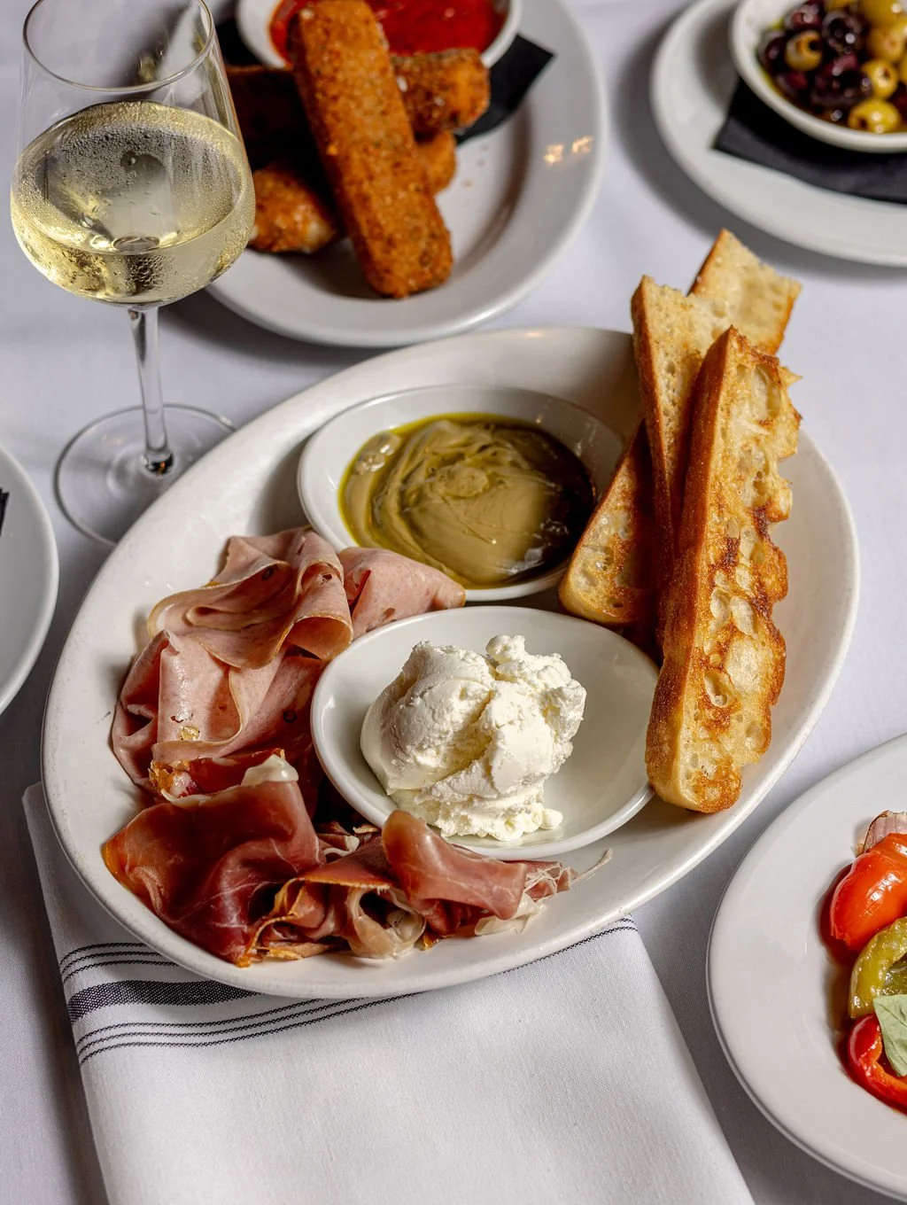 A platter of Italian antipasto with prosciutto, melted mozzarella, toasted bread slices, olive oil, and a small dish of honey, served on a white tablecloth. Additionally, a glass of white wine, fried mozzarella sticks with marinara sauce, and a bowl of mixed olives are visible.