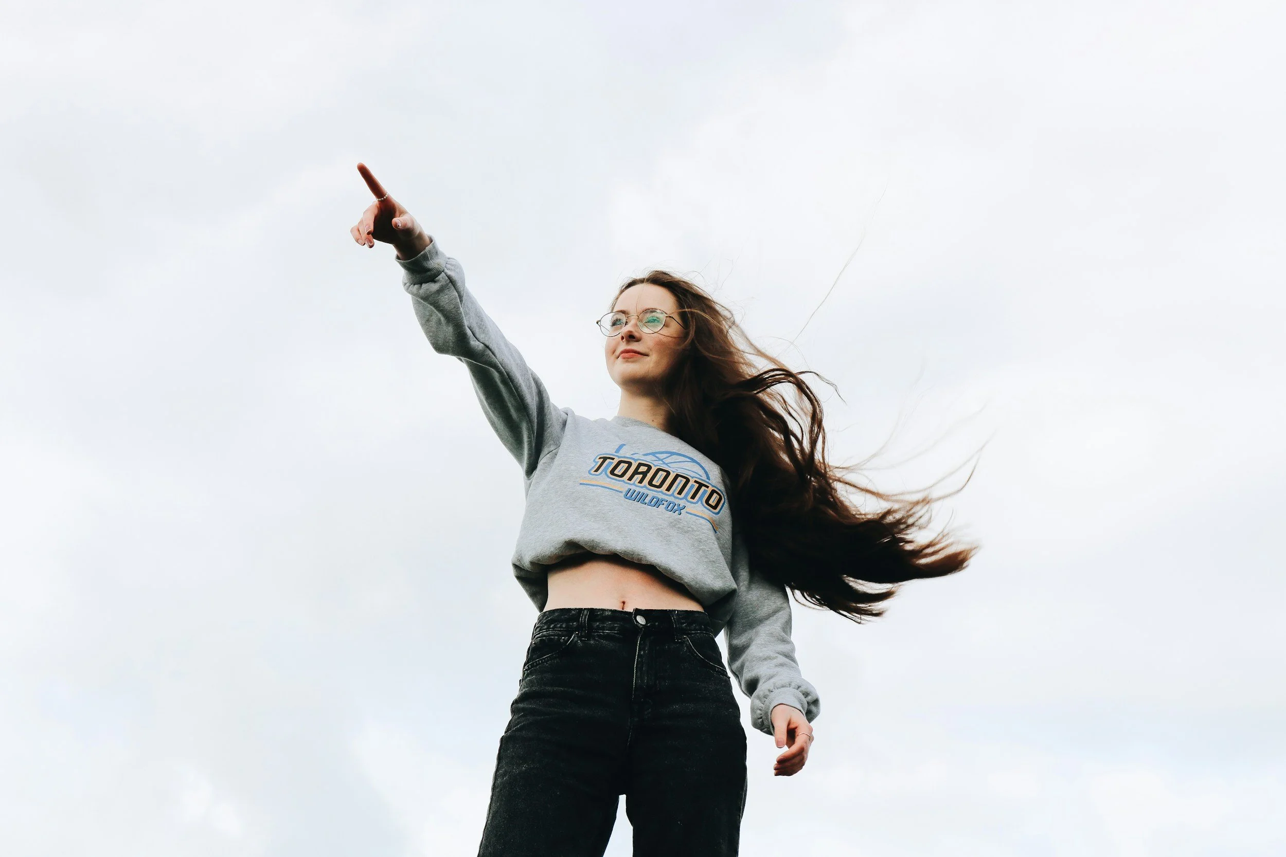 A young woman with long brown hair, glasses, and a gray Toronto sweatshirt, stands outdoors against a cloudy sky, pointing upward with her right hand.