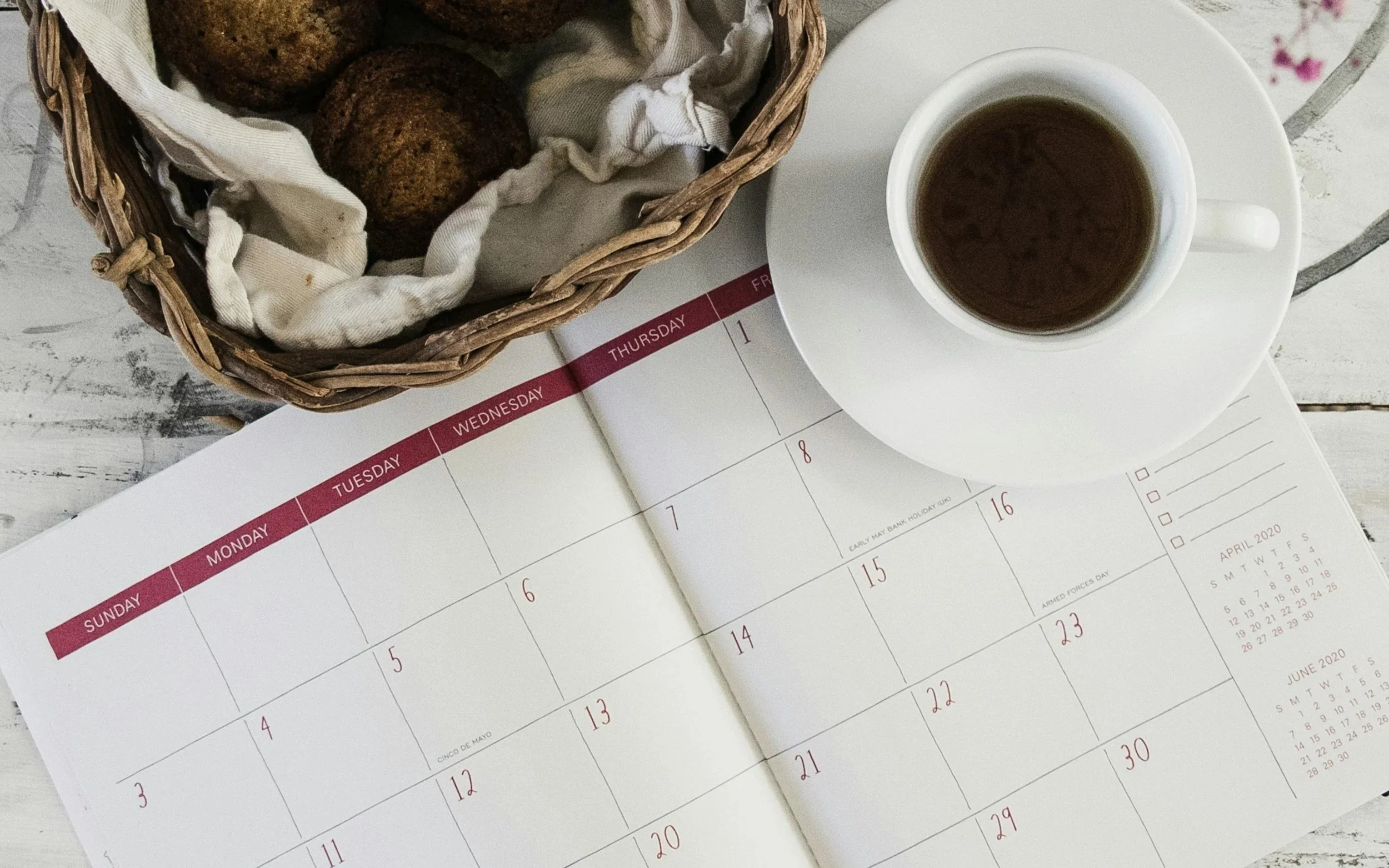A top-down view of a calendar opened to the month of April 2020, a cup of black coffee on a saucer, and a basket of cookies on a white wooden surface.
