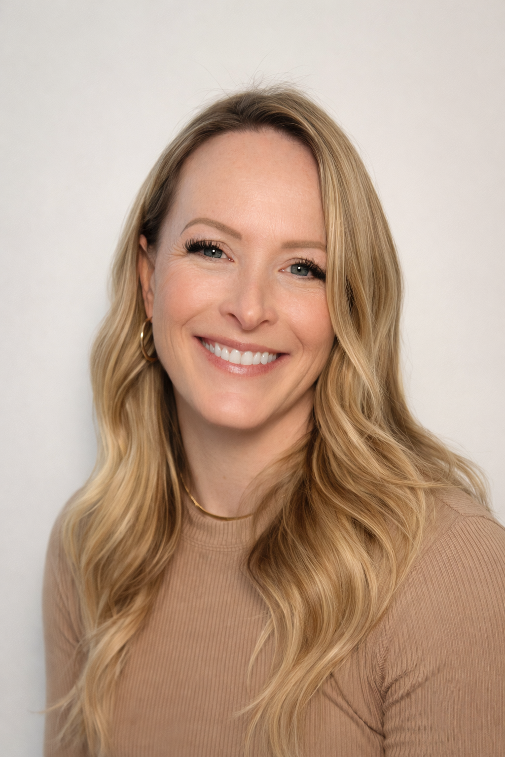 A woman with long, wavy blonde hair smiling at the camera, wearing a beige top and a gold necklace, against a plain white background.