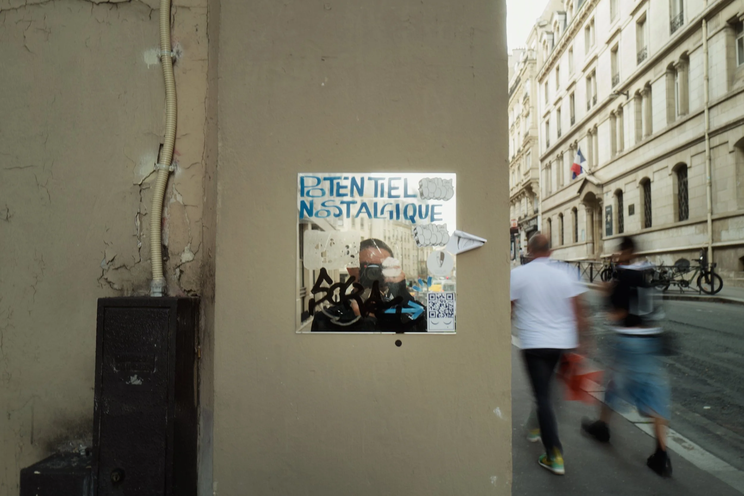 Street scene with a beige wall, graffiti, and a mirror with street reflections, showing three people walking past, in an urban area with classic European architecture.